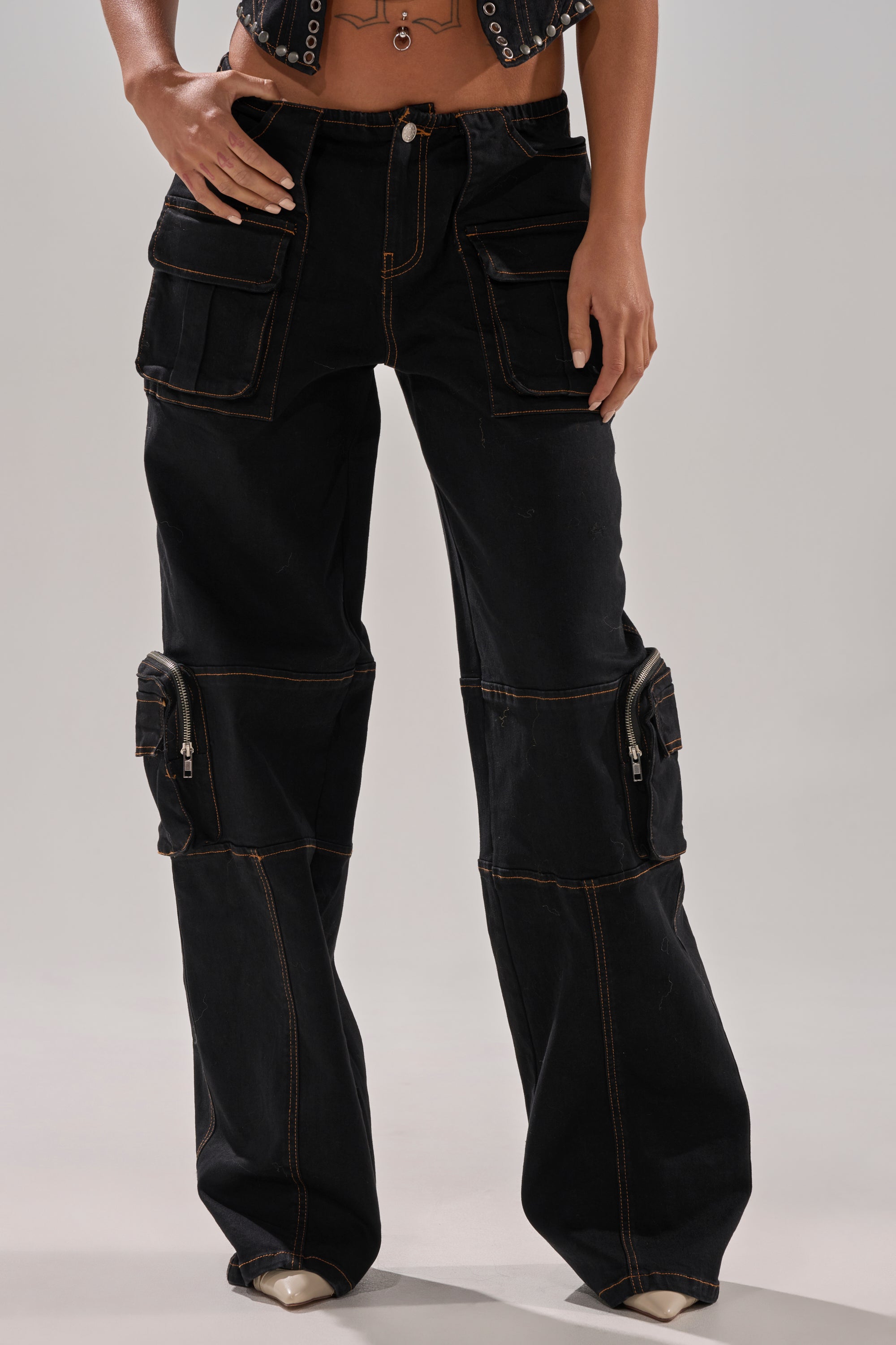 A model wears the FEELING FRISKY CARGO DENIM PANT—black, wide-leg cargo jeans with multiple large front pockets and contrast stitching—shown midriff to feet, standing against a plain background with hands on the pants.