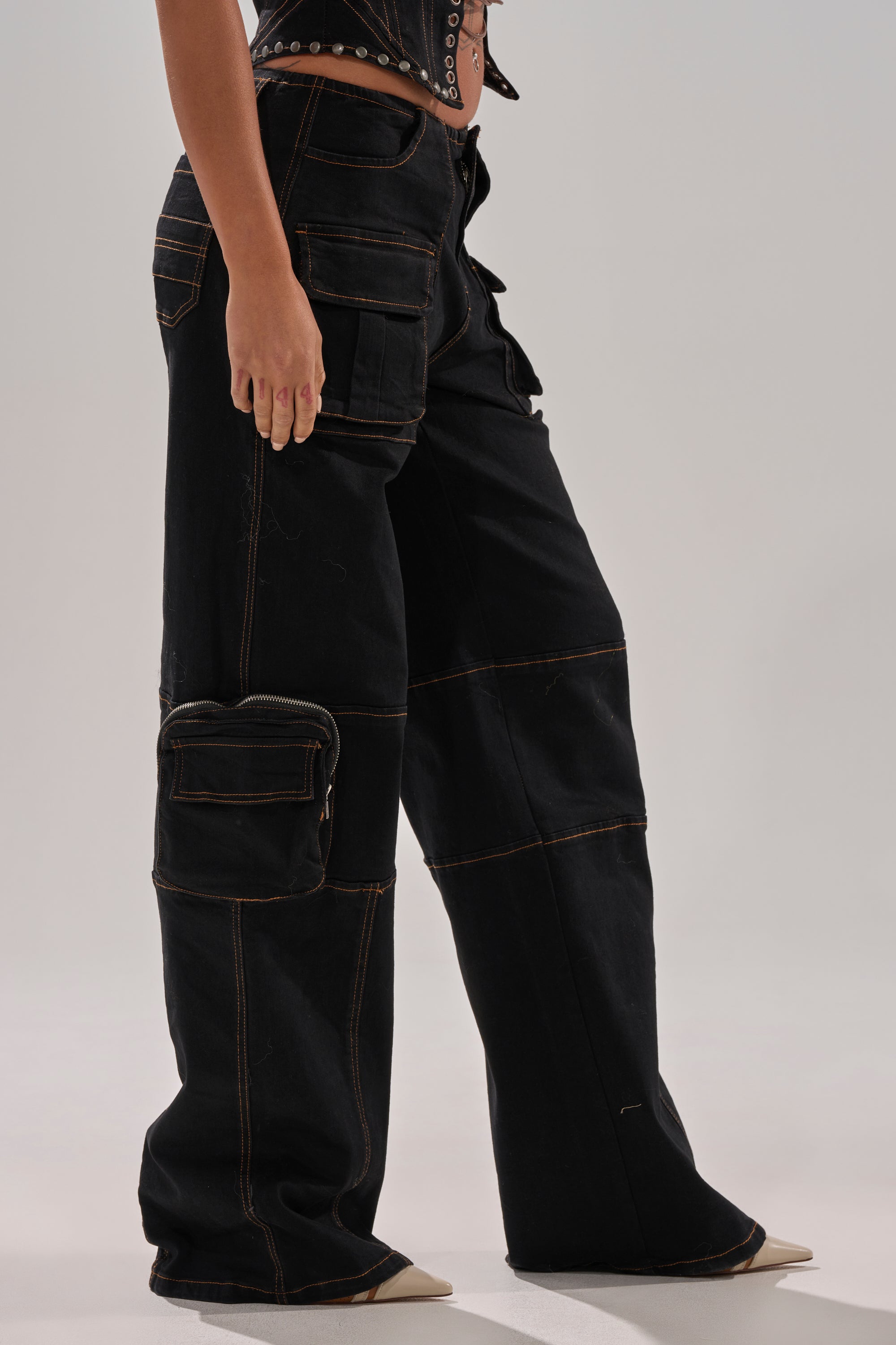 Someone stands against a plain light background wearing FEELING FRISKY CARGO DENIM PANT in loose-fitting black denim with a wide-leg silhouette, brown stitching, large cargo pockets, and beige pointed-toe shoes.
