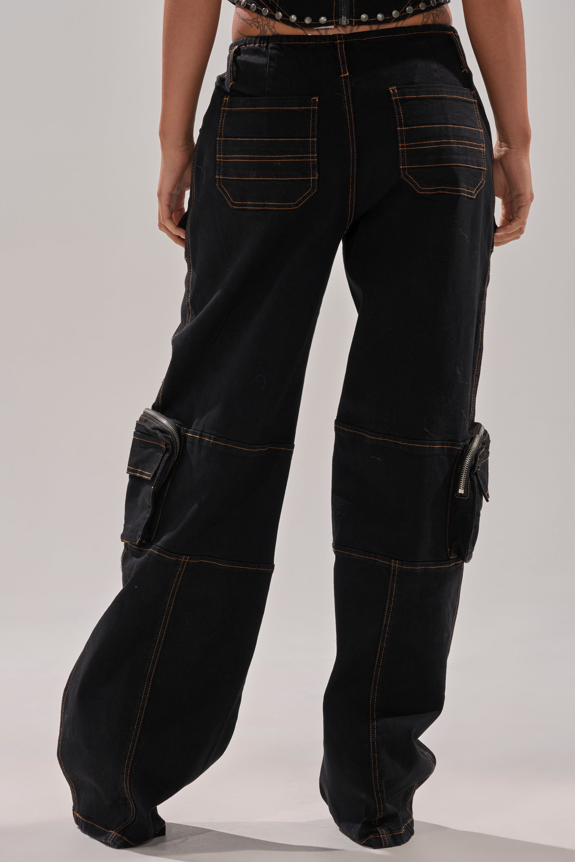 A person shown from the back wears the FEELING FRISKY CARGO DENIM PANT—loose-fitting black denim with a wide-leg silhouette, contrast stitching, and oversized back and side pockets—posed against a plain background.