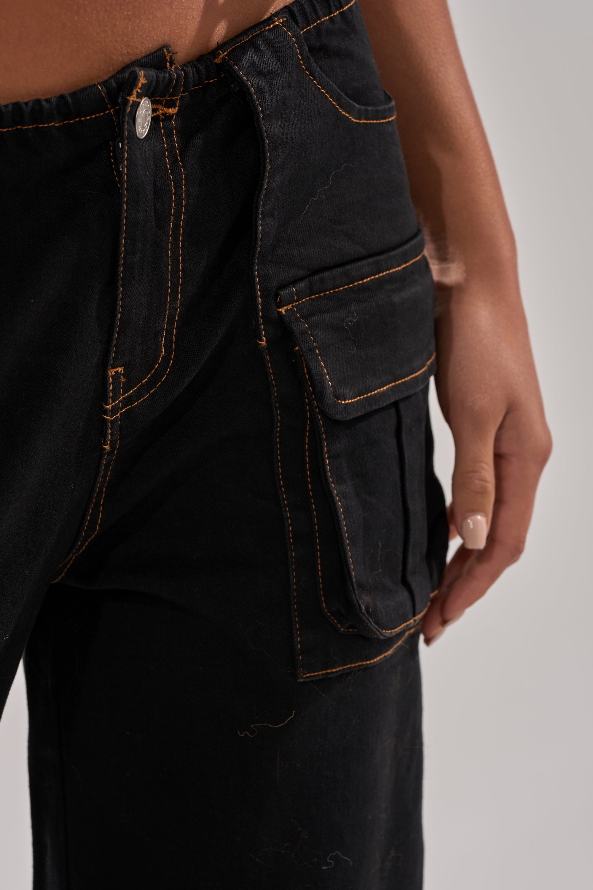 A close-up of a person wearing the FEELING FRISKY CARGO DENIM PANT, featuring black denim, orange stitching, a wide-leg fit, and a standout large side pocket as their manicured hand with light polish rests beside it.
