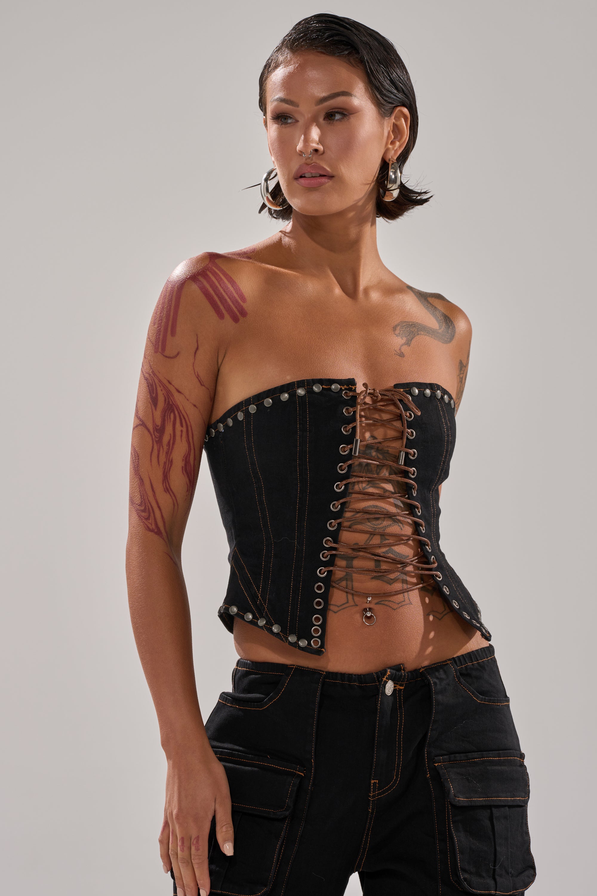 A woman in the FEELING FRISKY LACE UP CORSET strapless black top with matching pants poses with her hand on her hip; she has short, wet-look hair, visible arm and chest tattoos, and wears hoop earrings.