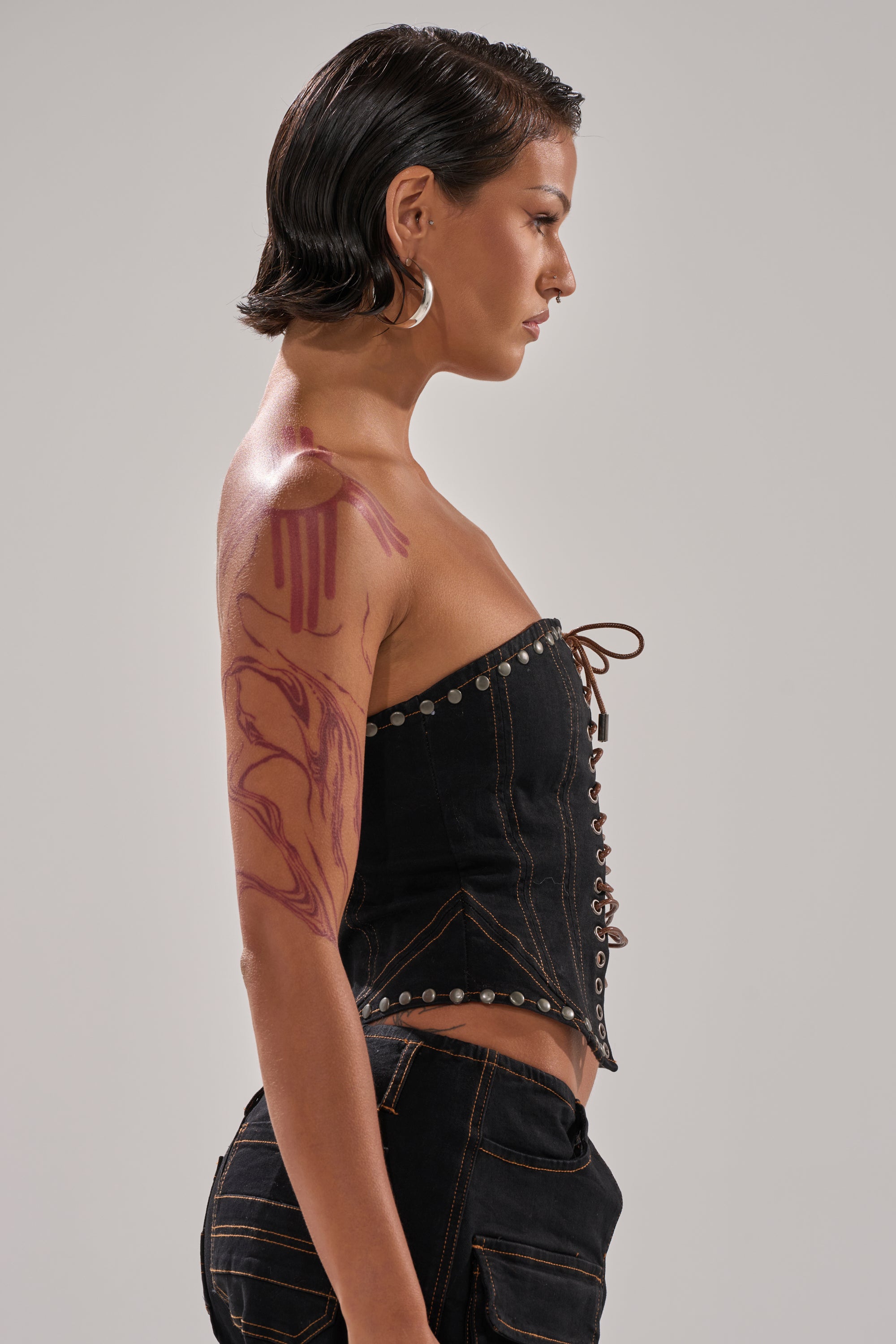 A person with short, slicked-back hair wears the FEELING FRISKY LACE UP CORSET in black with matching pants, revealing a maroon abstract arm design, and stands in profile against a plain background.
