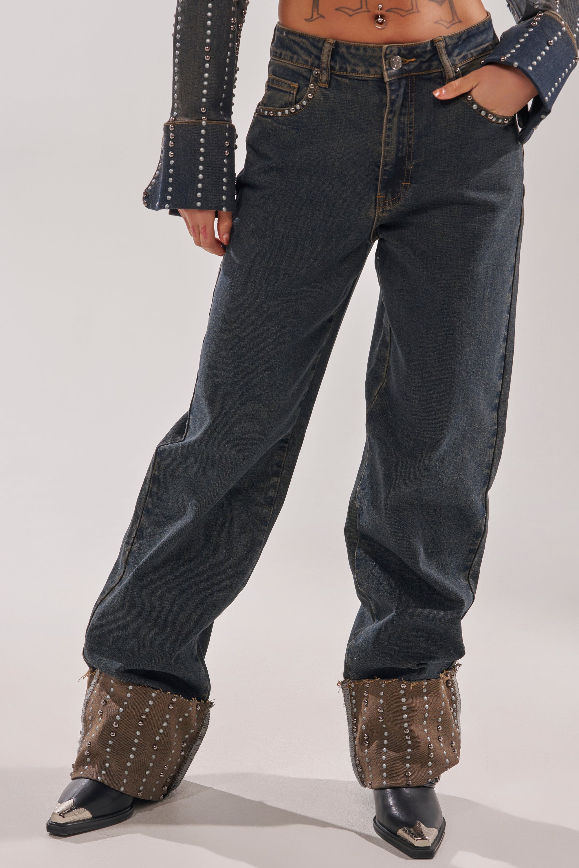 A person models the VANJA STUDDED WIDE LEG DENIM with metal-studded cuffs, a matching flared-sleeve denim top, and pointed-toe black boots. Only the torso and legs are shown. 2