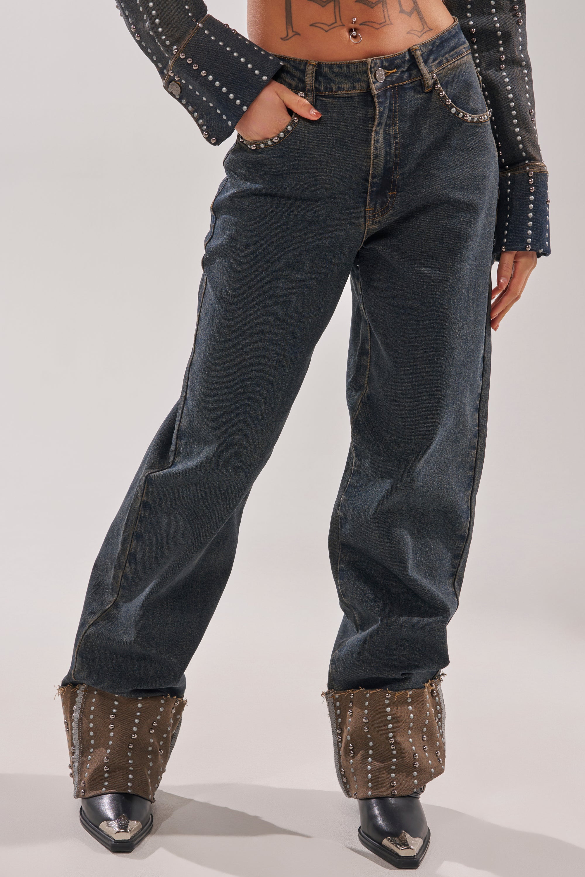 Wearing the VANJA STUDDED WIDE LEG DENIM with one hand in the pocket, a studded dark top, and brown studded leg warmers over pointed black shoes, their tattooed midriff is visible. 0