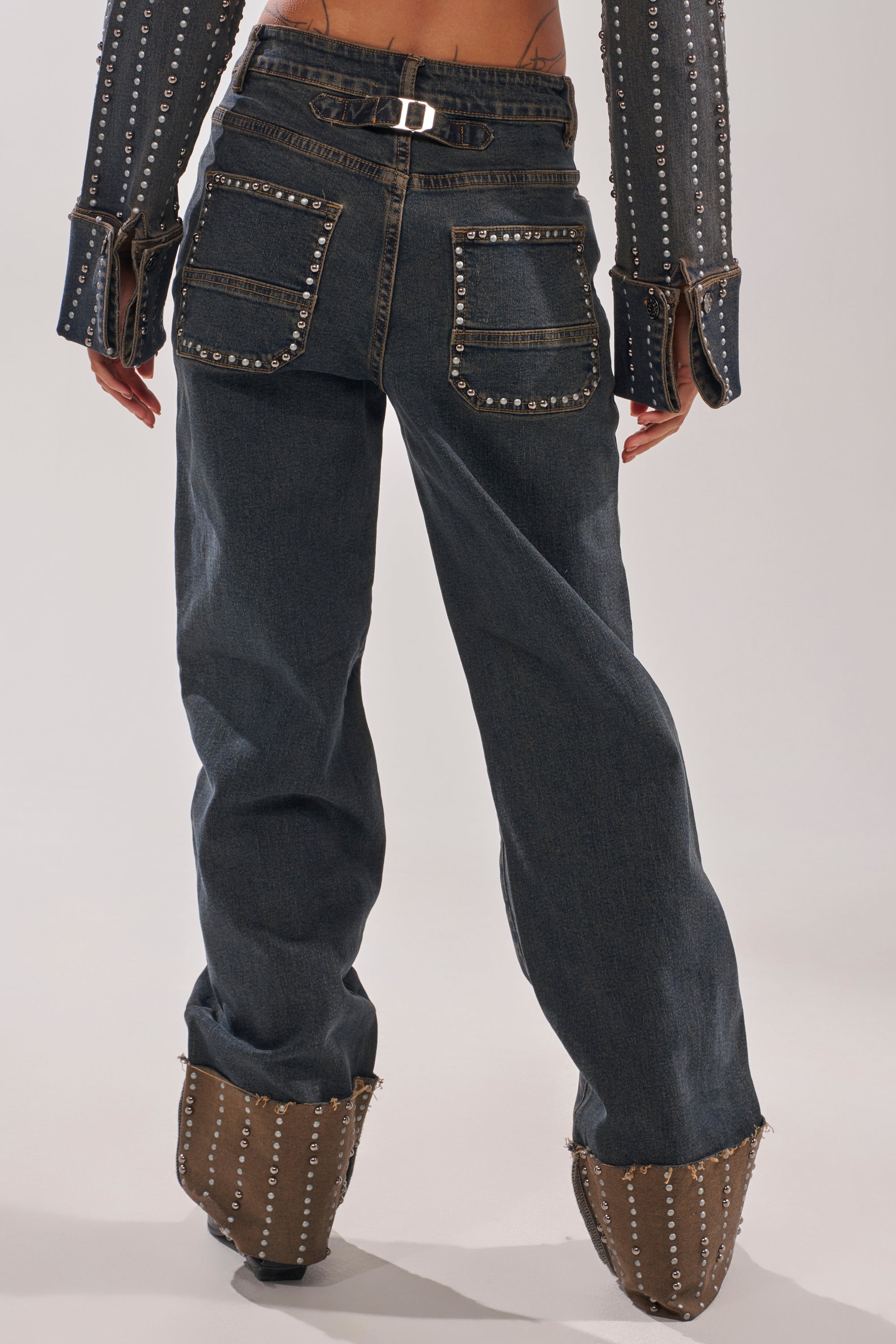 A person wearing VANJA STUDDED WIDE LEG DENIM jeans with large back pockets, metal details, and matching studded cuffs and shoes stands against a plain light background, photographed from behind. 4