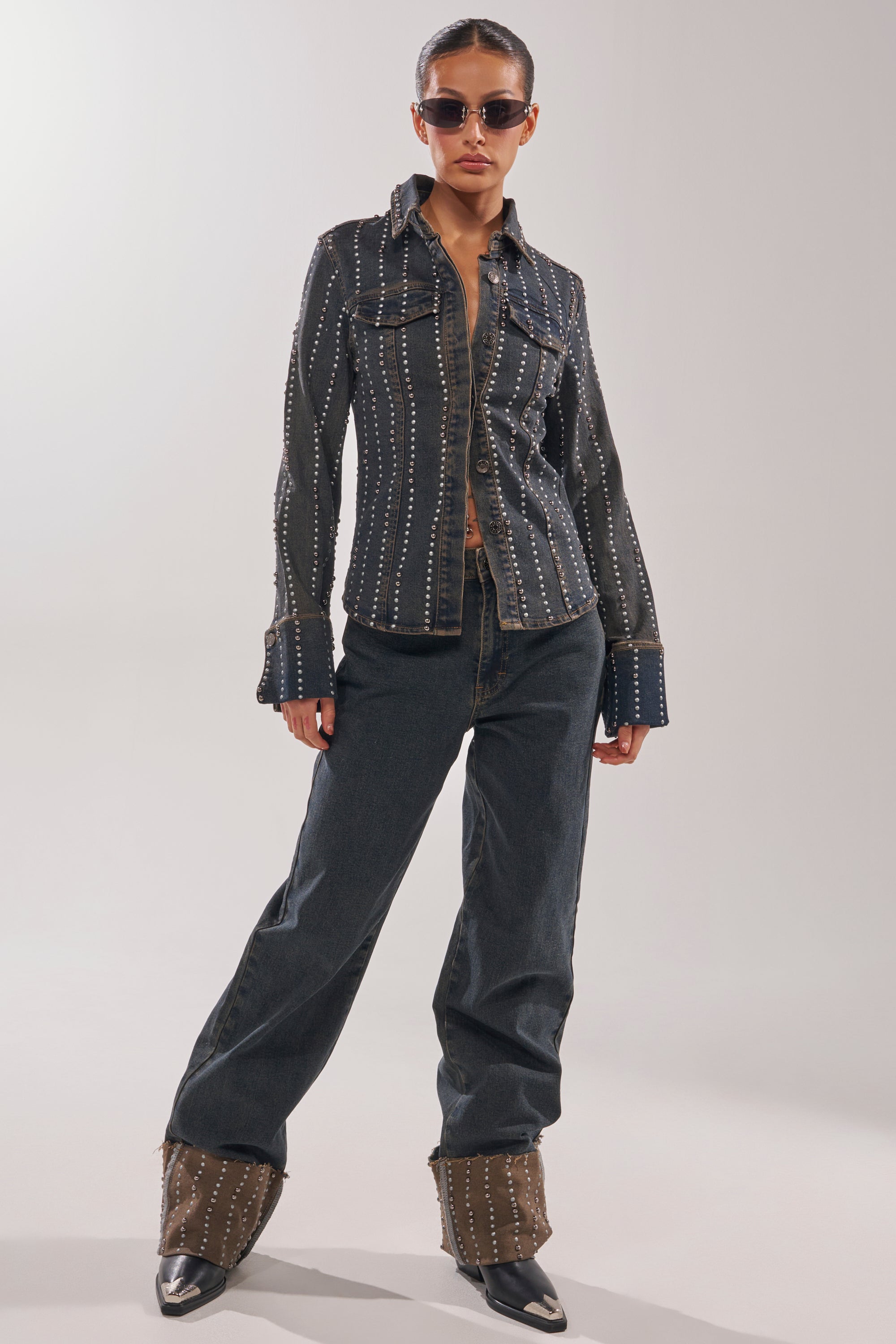 A person wearing sunglasses, an AKIRA Label dark studded button-up shirt with flared cuffs, VANJA STUDDED WIDE LEG DENIM jeans, and pointed boots stands confidently against a plain background. 5