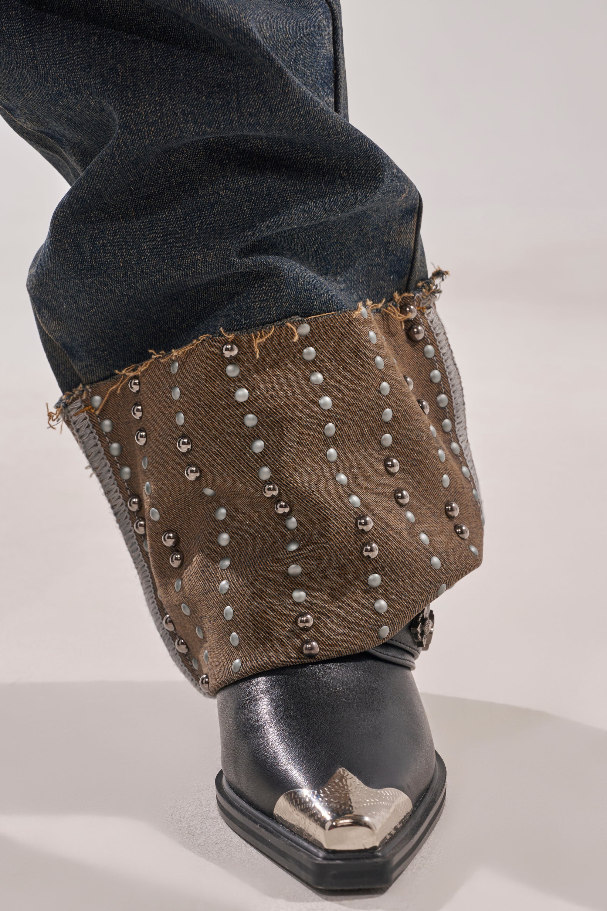 A close-up of a foot in a black pointed cowboy boot with a silver toe cap, partially covered by frayed brown fabric and vertical silver studs, styled over the VANJA STUDDED WIDE LEG DENIM. 7