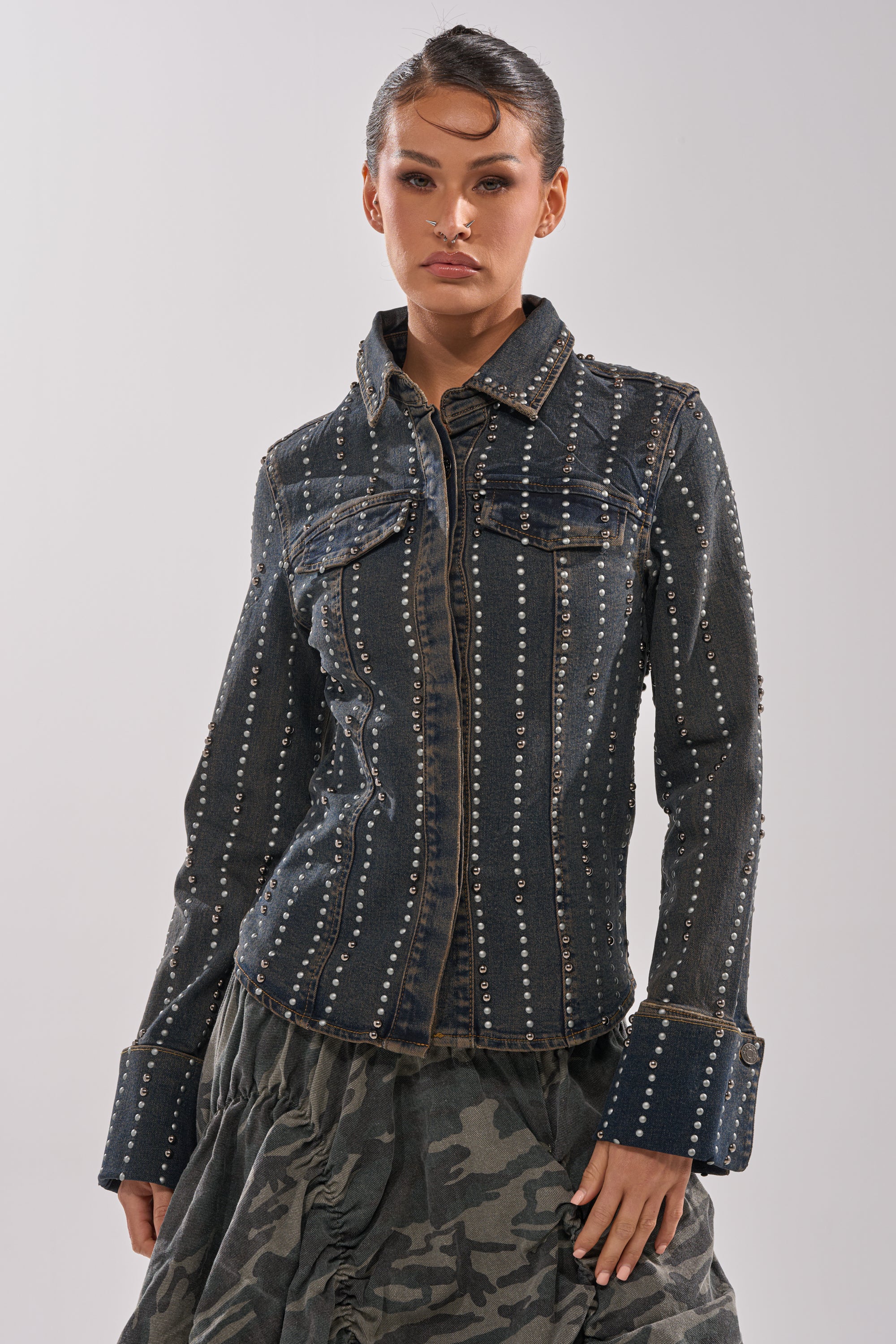 A woman poses against a plain light background wearing the VANJA STUDDED DENIM BUTTON DOWN with vertical studs, large cuffs, and a ruffled camouflage skirt.