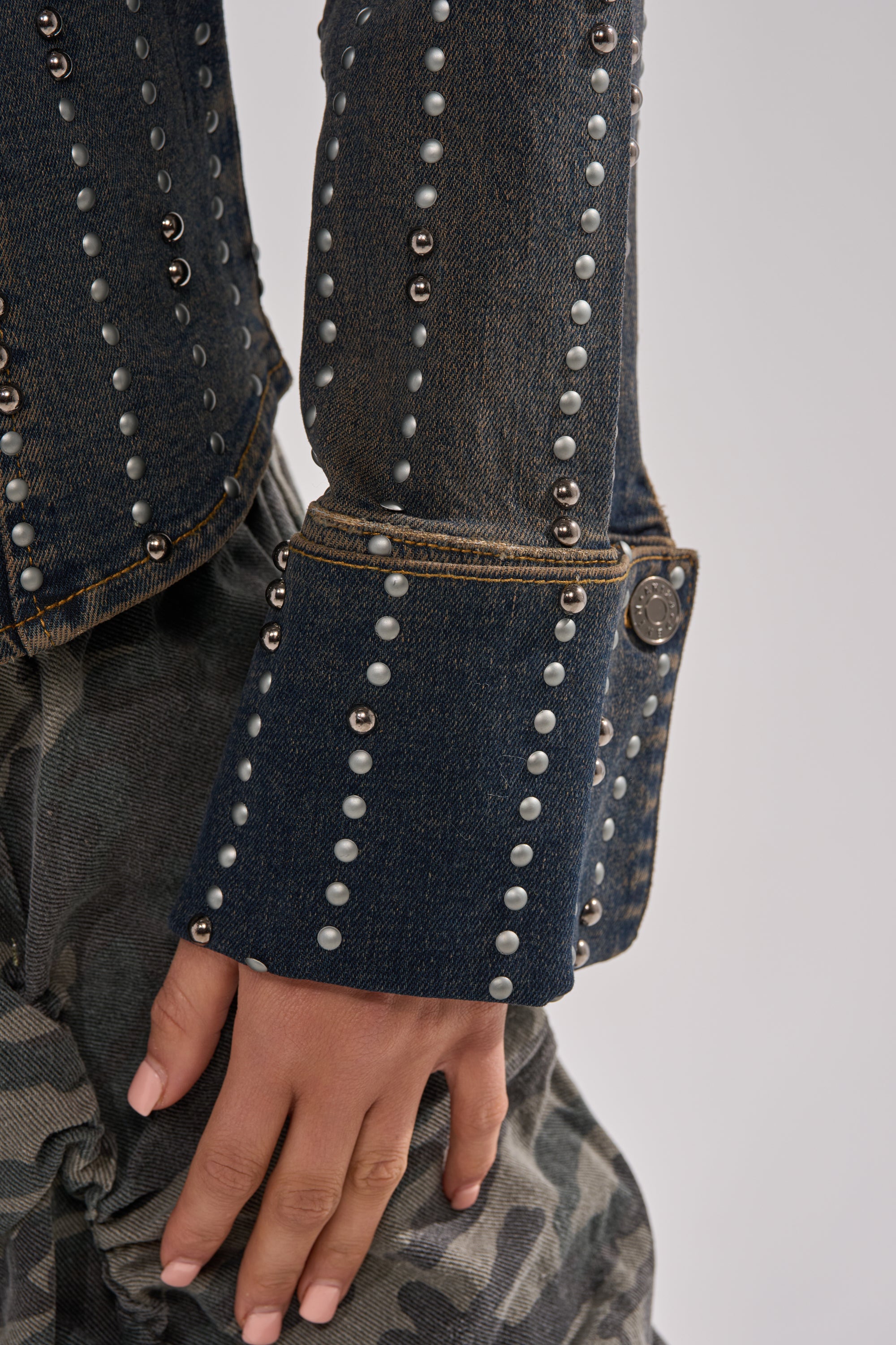 Close-up of a person in the VANJA STUDDED DENIM BUTTON DOWN with pearl and metal embellishments, wide cuffs, and camo pants. Their hand, near their hip, showcases neatly manicured nails.