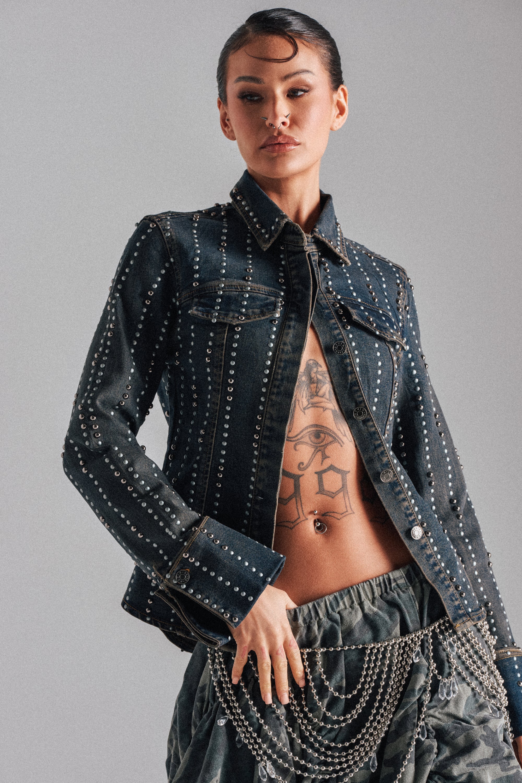Confidently posing in the VANJA STUDDED DENIM BUTTON DOWN left open to show a chest tattoo and belly button ring, they complete the look with dark pants featuring draped silver chains.