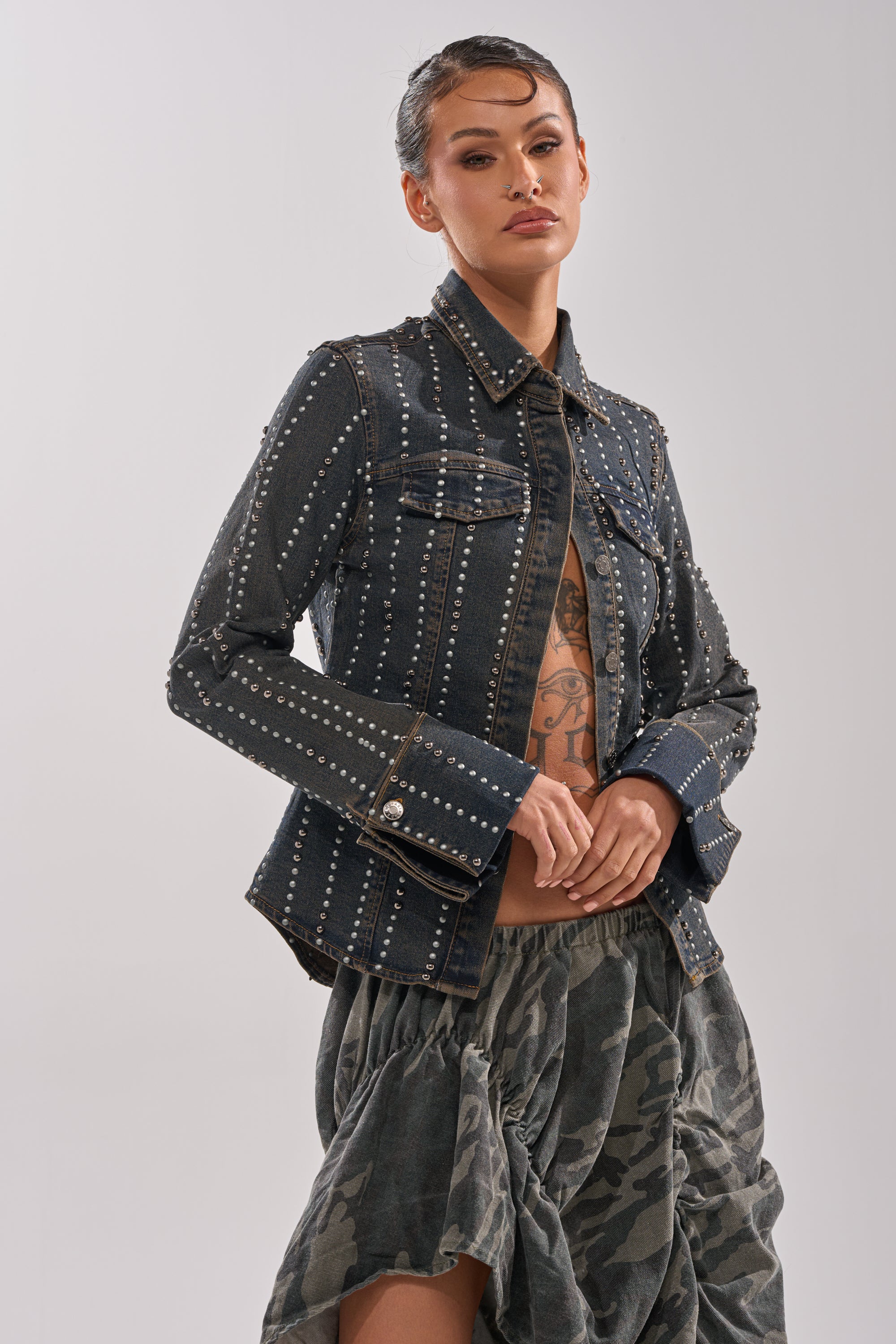 A woman poses confidently against a plain background in the VANJA STUDDED DENIM BUTTON DOWN over a midriff-baring top and a voluminous camo skirt.