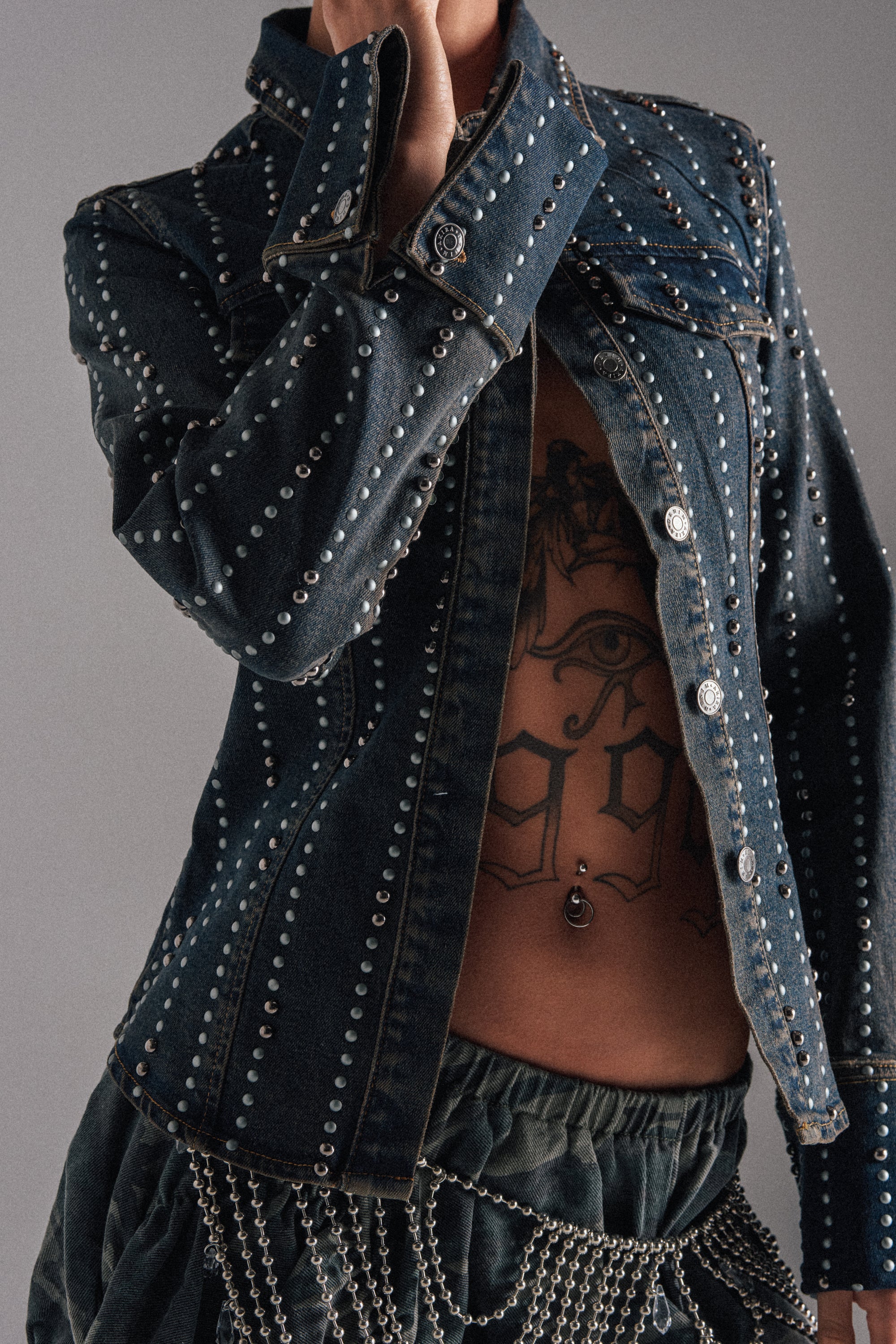 Wearing the VANJA STUDDED DENIM BUTTON DOWN and green chain-detailed pants, a person reveals their tattooed torso and belly button piercing while lifting the jacket collar to partially hide their face.