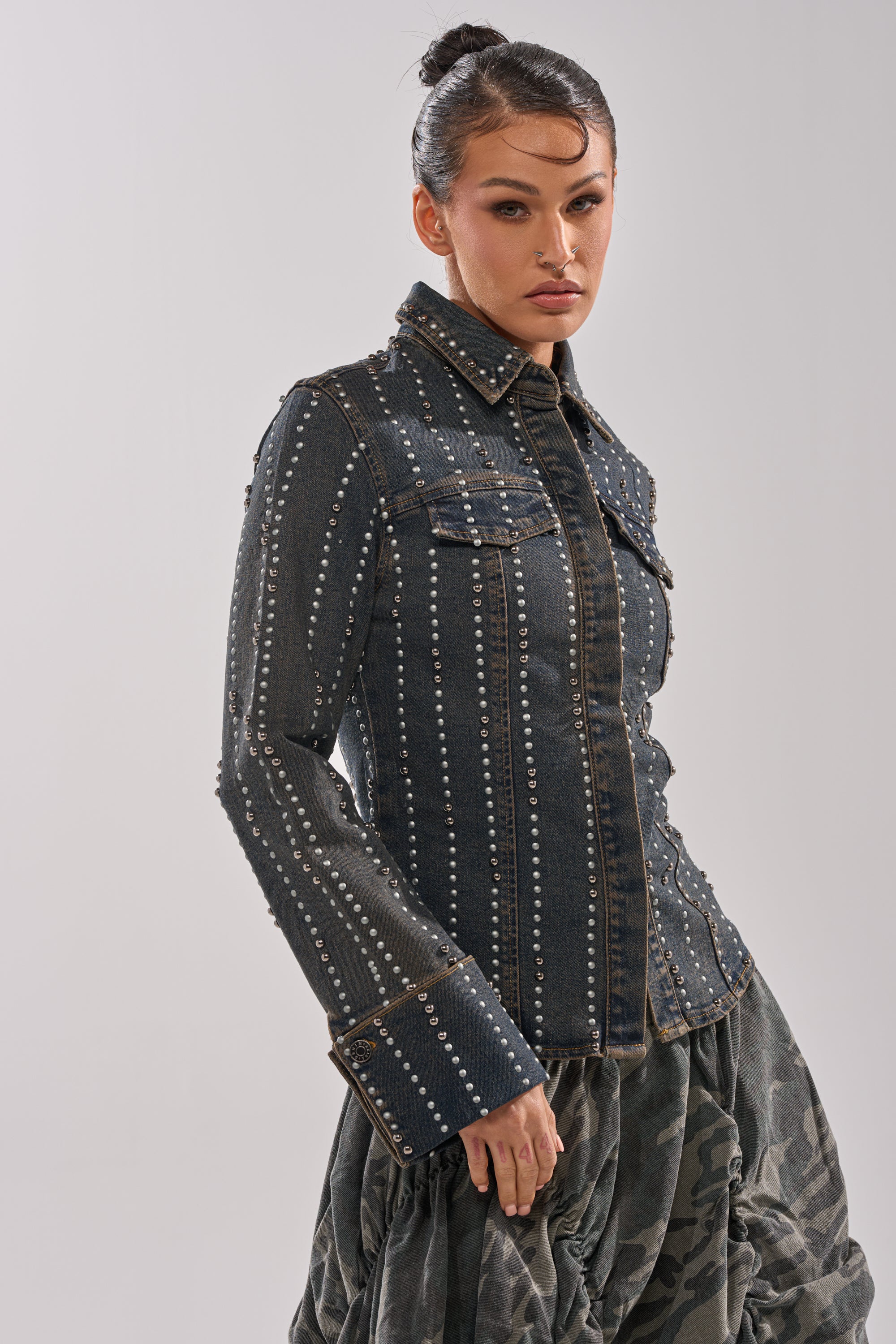 A woman with hair in a bun models the VANJA STUDDED DENIM BUTTON DOWN, styled with a gray, textured, voluminous skirt. She poses confidently against a light neutral background.