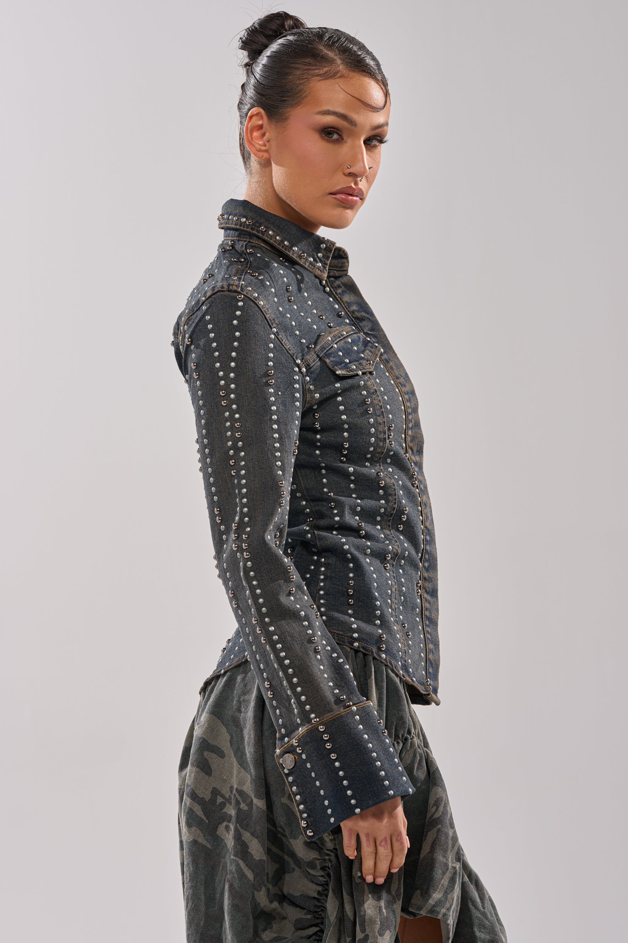 A woman with dark hair in a bun wears the VANJA STUDDED DENIM BUTTON DOWN and a patterned skirt, standing sideways and looking toward the camera against a plain light background.