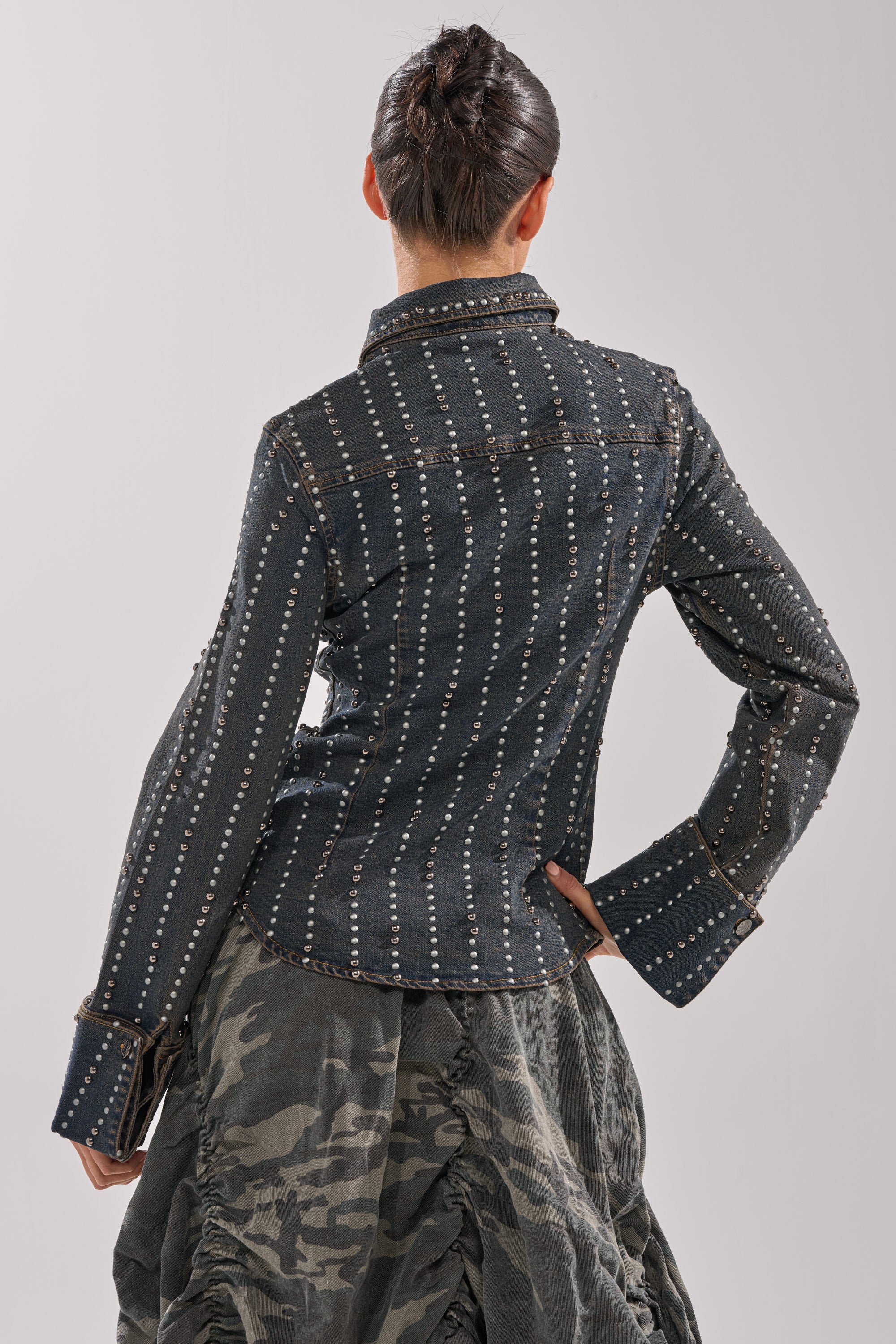A person stands with their back to the camera, wearing the VANJA STUDDED DENIM BUTTON DOWN with wide cuffs, paired with a voluminous, ruched camouflage skirt.