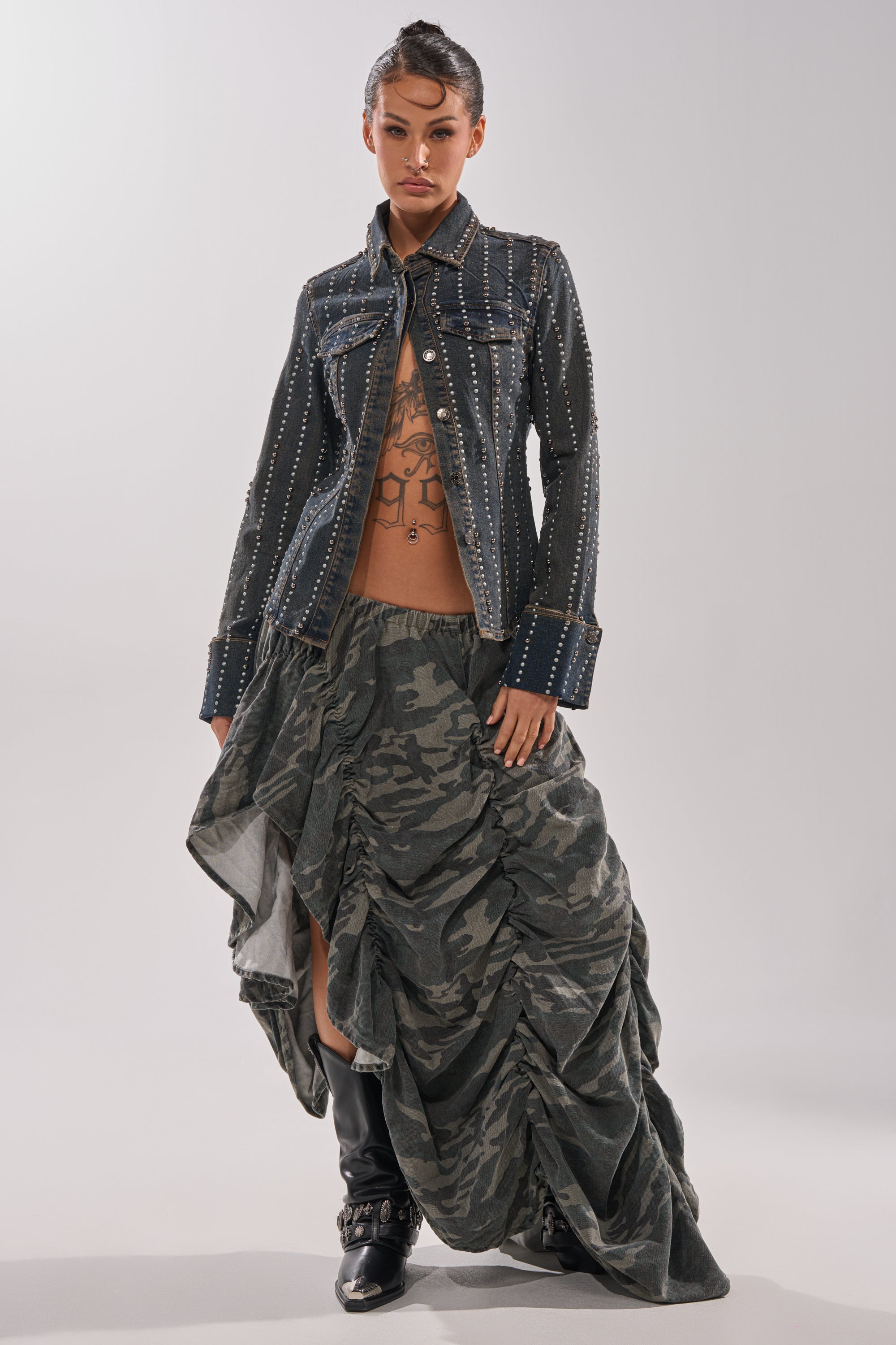 A woman poses in the VANJA STUDDED DENIM BUTTON DOWN, worn open to show her tattooed torso, styled with a ruched camo maxi skirt and black boots against a plain background.