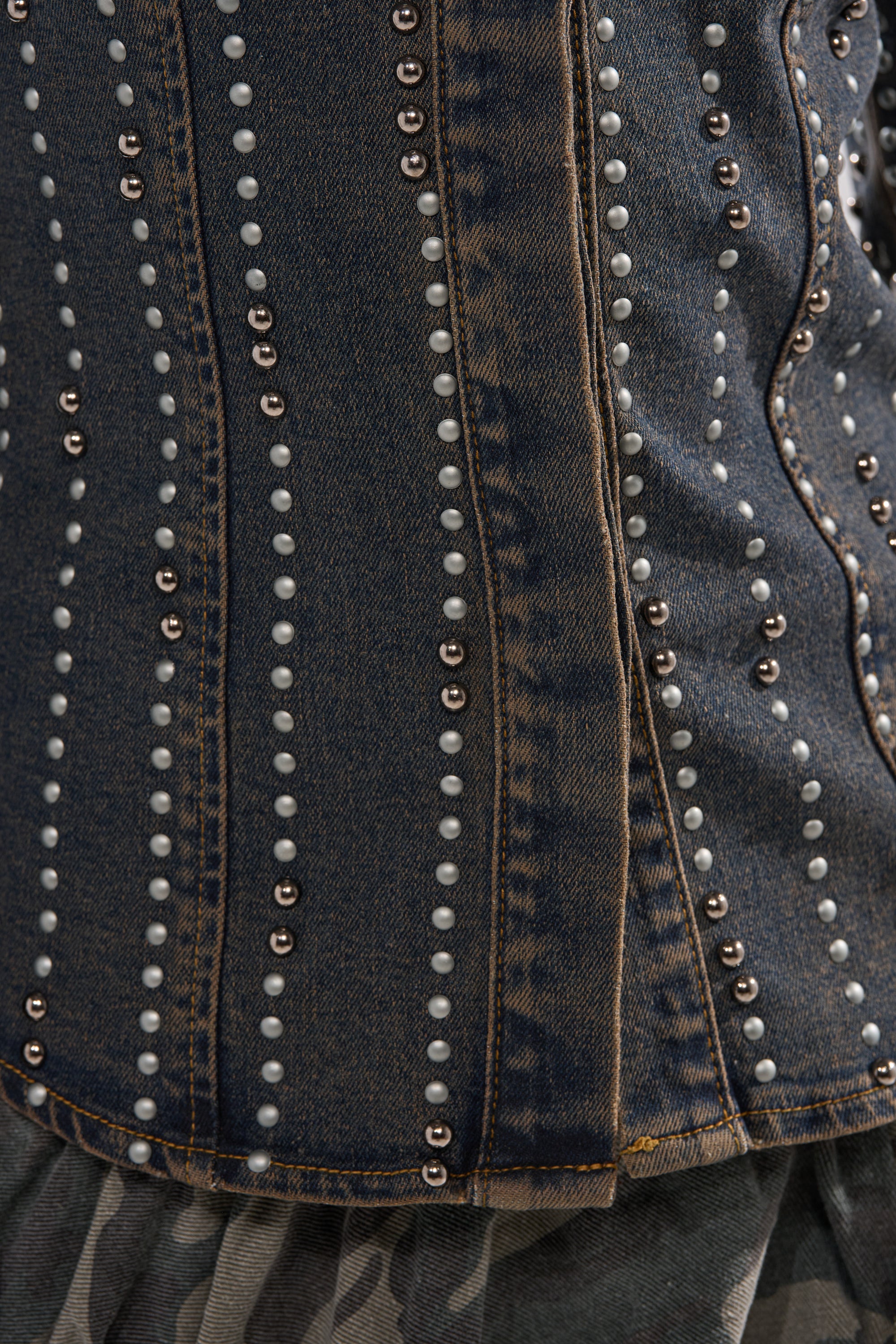 Close-up of the VANJA STUDDED DENIM BUTTON DOWN, showcasing blue denim with vertical white and metallic studs, detailed stitching, and a touch of grey striped fabric at the hem.