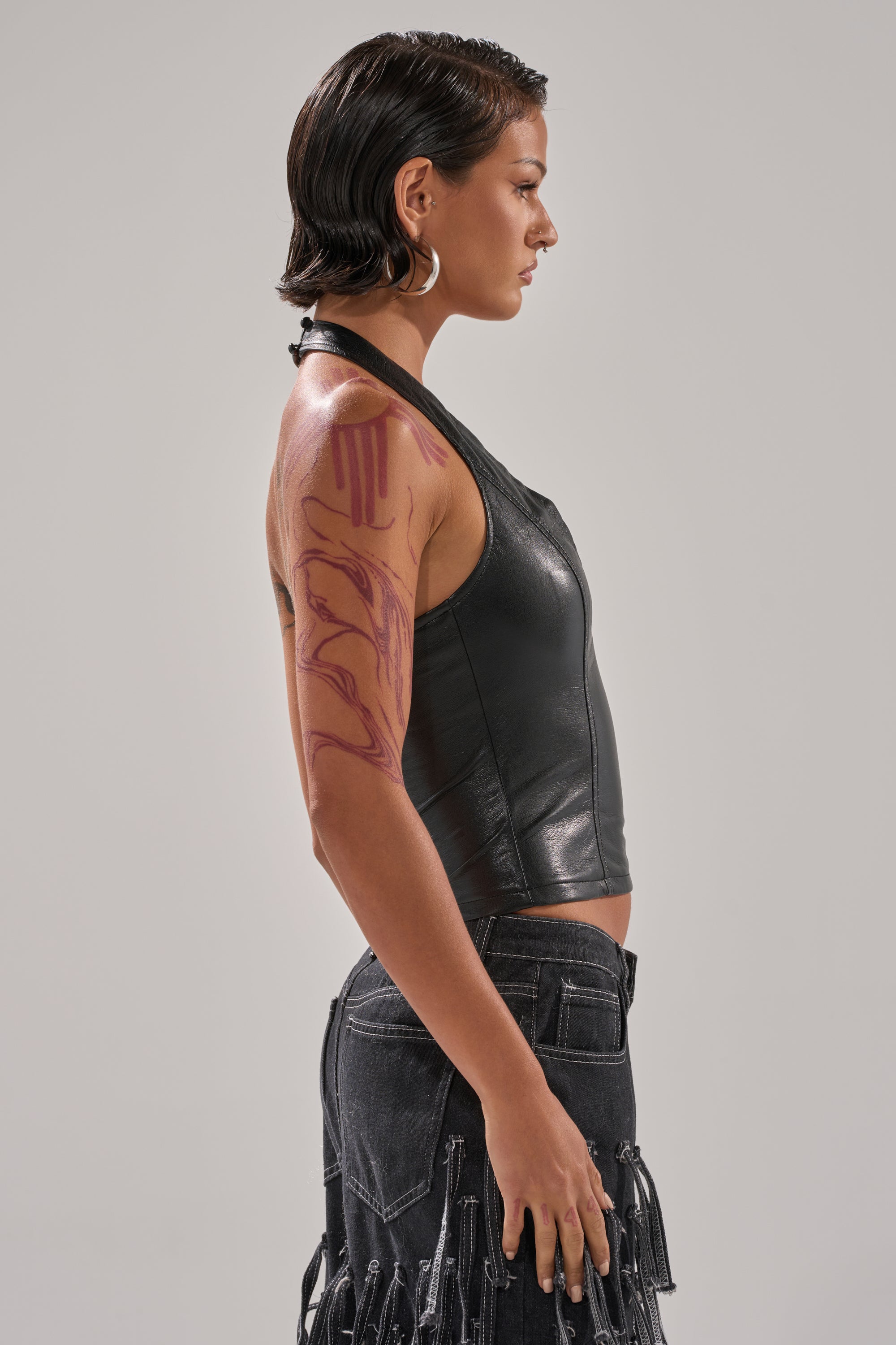 A woman with short, slicked-back hair wears the WITHOUT A DOUBT FAUX LEATHER HALTER NECK CORSET IN BLACK and a fringe skirt, accessorized with large hoop earrings and abstract red body art on her upper arm, posed in profile against a plain background.