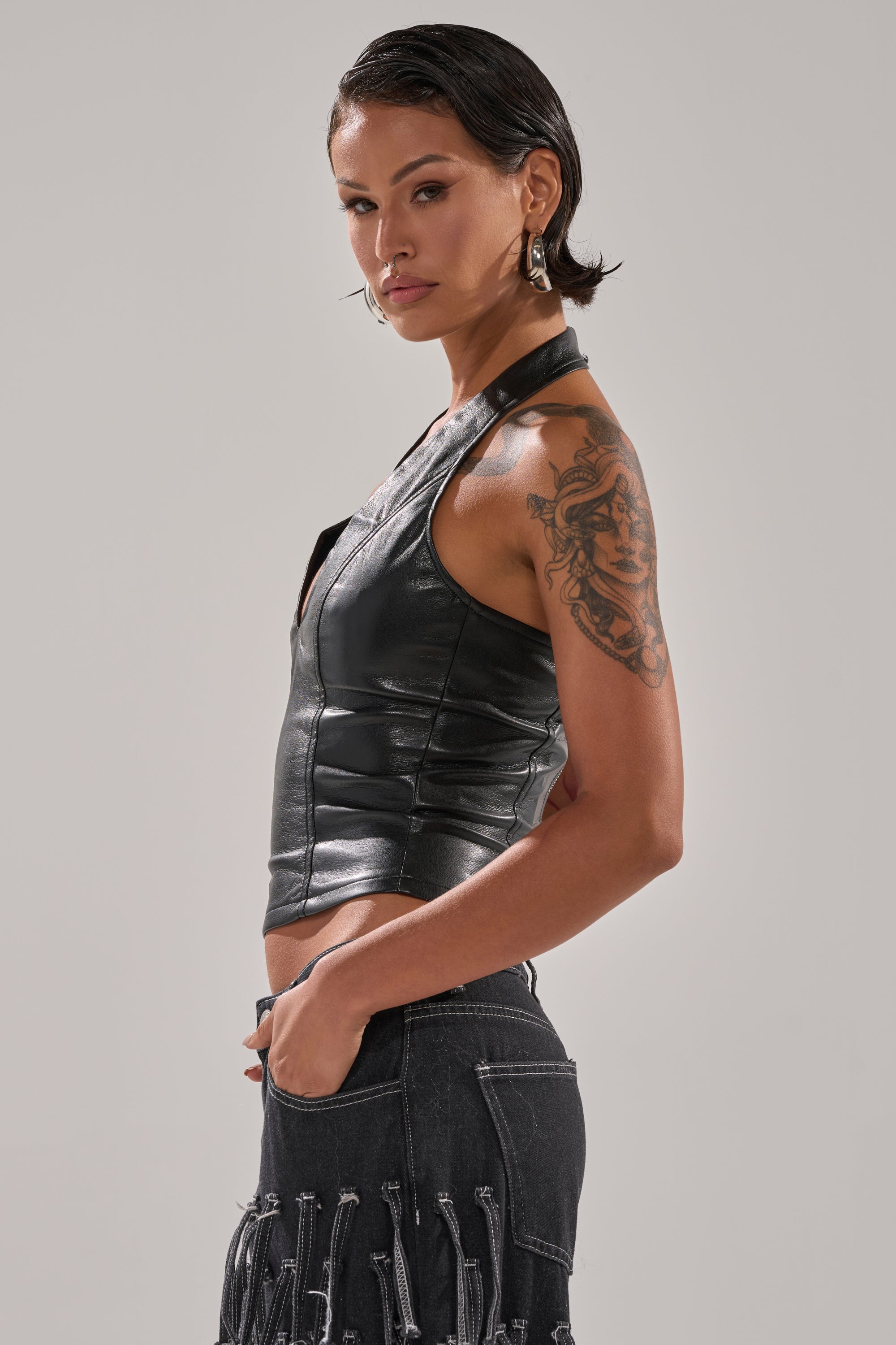 A woman with short dark hair wears the WITHOUT A DOUBT FAUX LEATHER HALTER NECK CORSET IN BLACK, paired with a fringed dark denim skirt and hoop earrings. She stands in profile, showing her upper arm tattoo against a plain background.