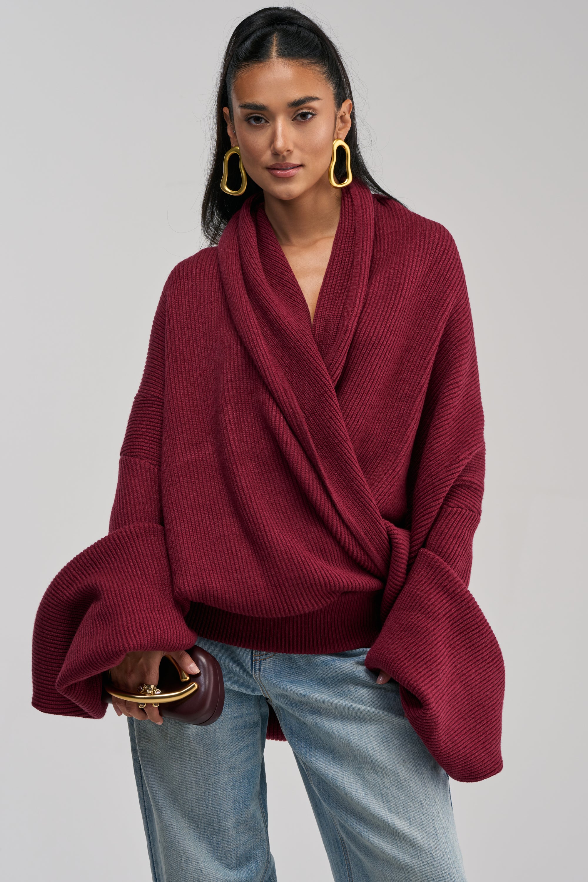 Wearing the CRAZY FOR YOU CROSS FRONT SWEATER IN BURGUNDY—a loose knit wrap style with an oversized shawl collar—a person stands in light blue jeans, holding a small burgundy clutch. Their long dark hair, gold earrings, and neutral expression are set against a plain background.