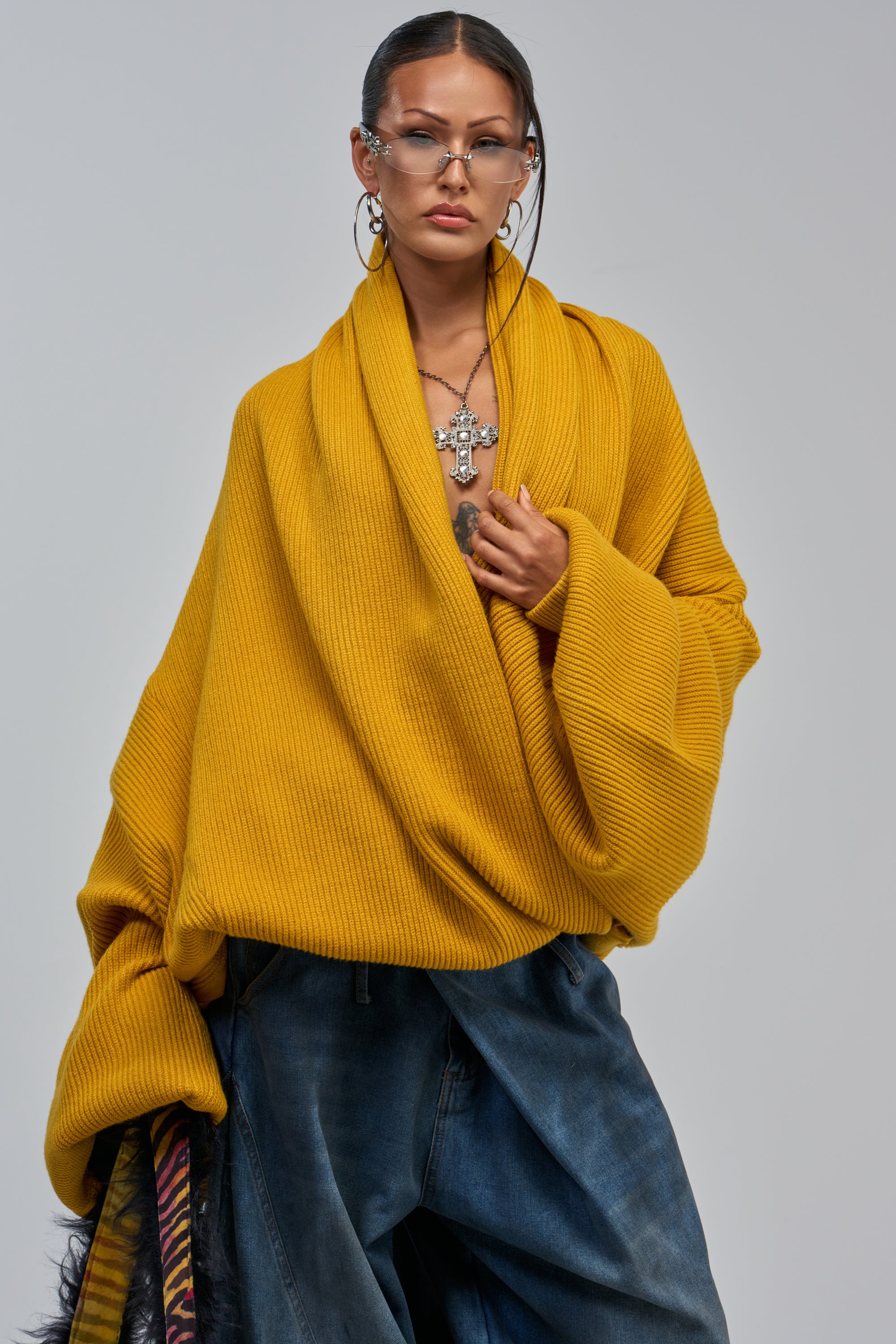 Wearing the CRAZY FOR YOU CROSS FRONT SWEATER IN MUSTARD, blue jeans, hoops, layered necklaces, and glasses, a person poses confidently against a plain background.
