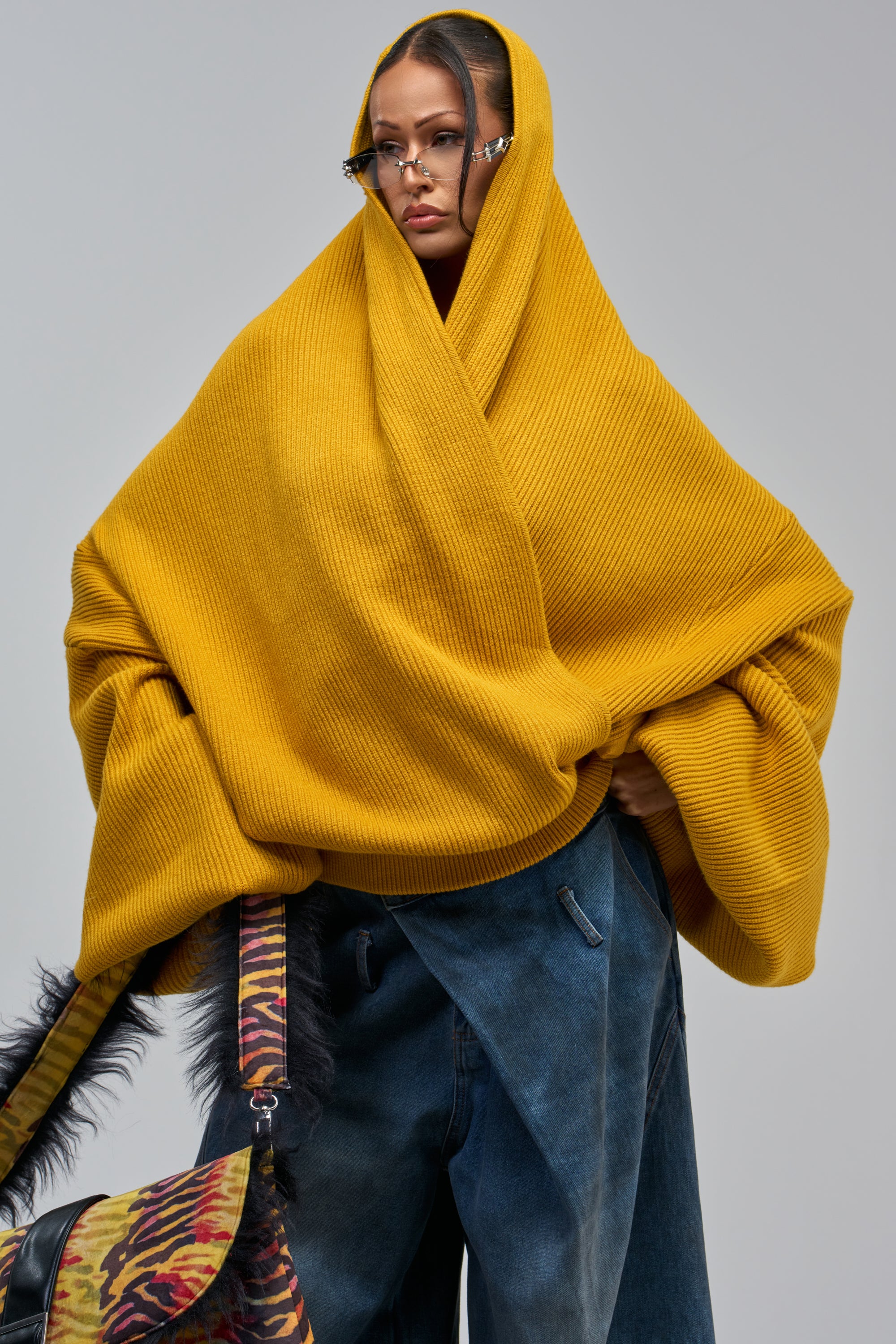 A person wears the CRAZY FOR YOU CROSS FRONT SWEATER IN MUSTARD, loose blue jeans, small sunglasses, and carries a colorful animal print bag with black feather details.
