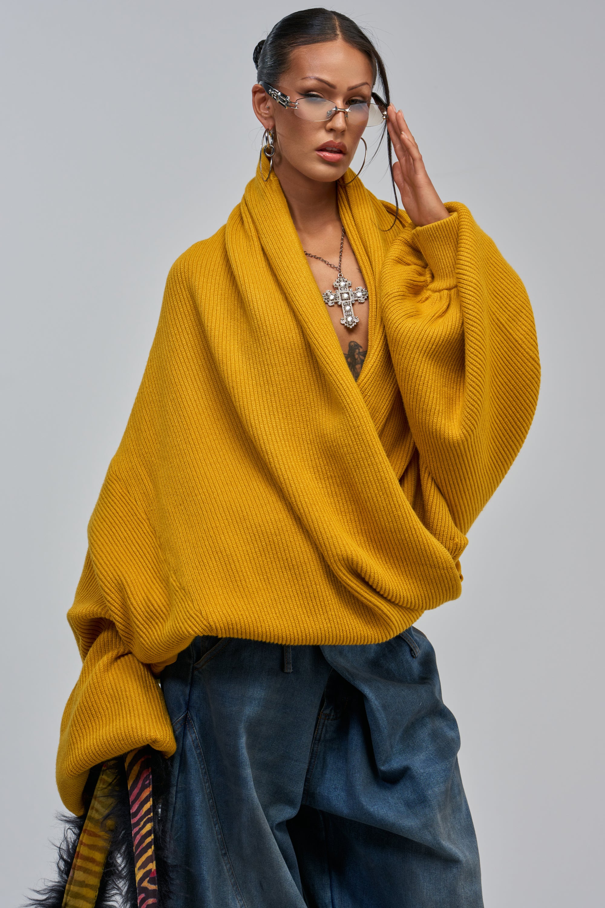 Model wears the CRAZY FOR YOU CROSS FRONT SWEATER IN MUSTARD with wide-leg jeans, statement earrings, and layered cross necklaces, posing with one hand on their face against a neutral background.