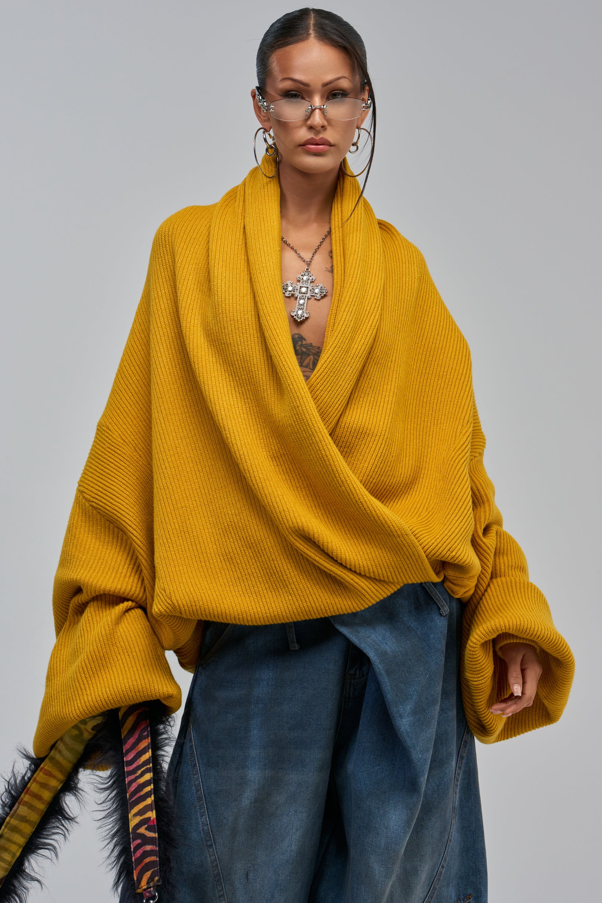 A woman stands confidently in the CRAZY FOR YOU CROSS FRONT SWEATER IN MUSTARD and wide-leg jeans, accessorized with glasses, hoop earrings, layered cross necklaces, and a colorful striped bag with fur trim.