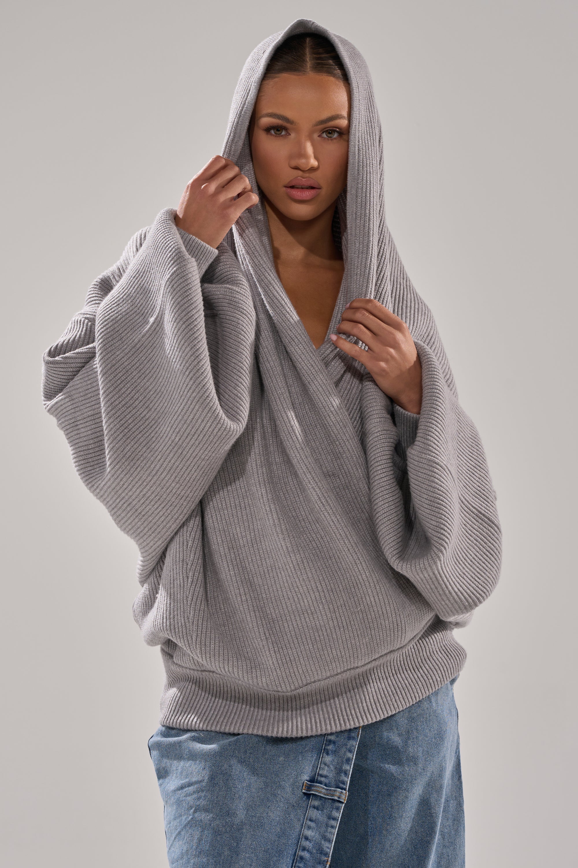 A woman wears the CRAZY FOR YOU CROSS FRONT SWEATER, a loose heather grey knit with a hood, paired with blue jeans. She stands against a plain light gray background, holding the hood and looking at the camera. 0