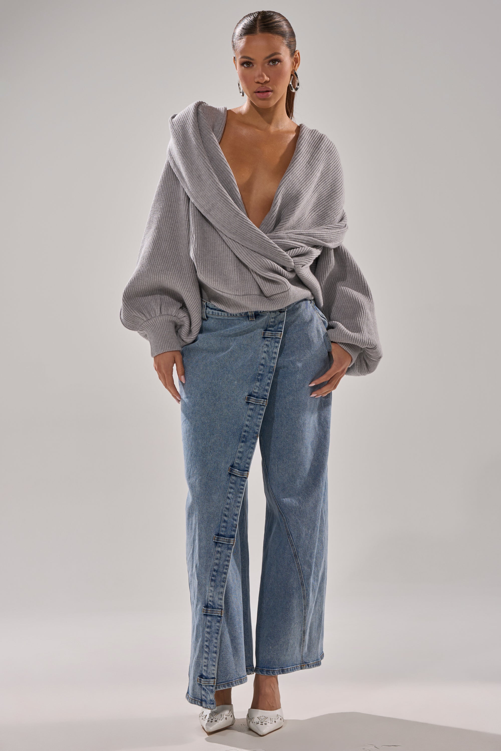 A woman poses against a plain background wearing the CRAZY FOR YOU CROSS FRONT SWEATER in heather grey, featuring balloon sleeves and a deep V-neck, paired with high-waisted wide-leg blue jeans and white pointed heels. 9