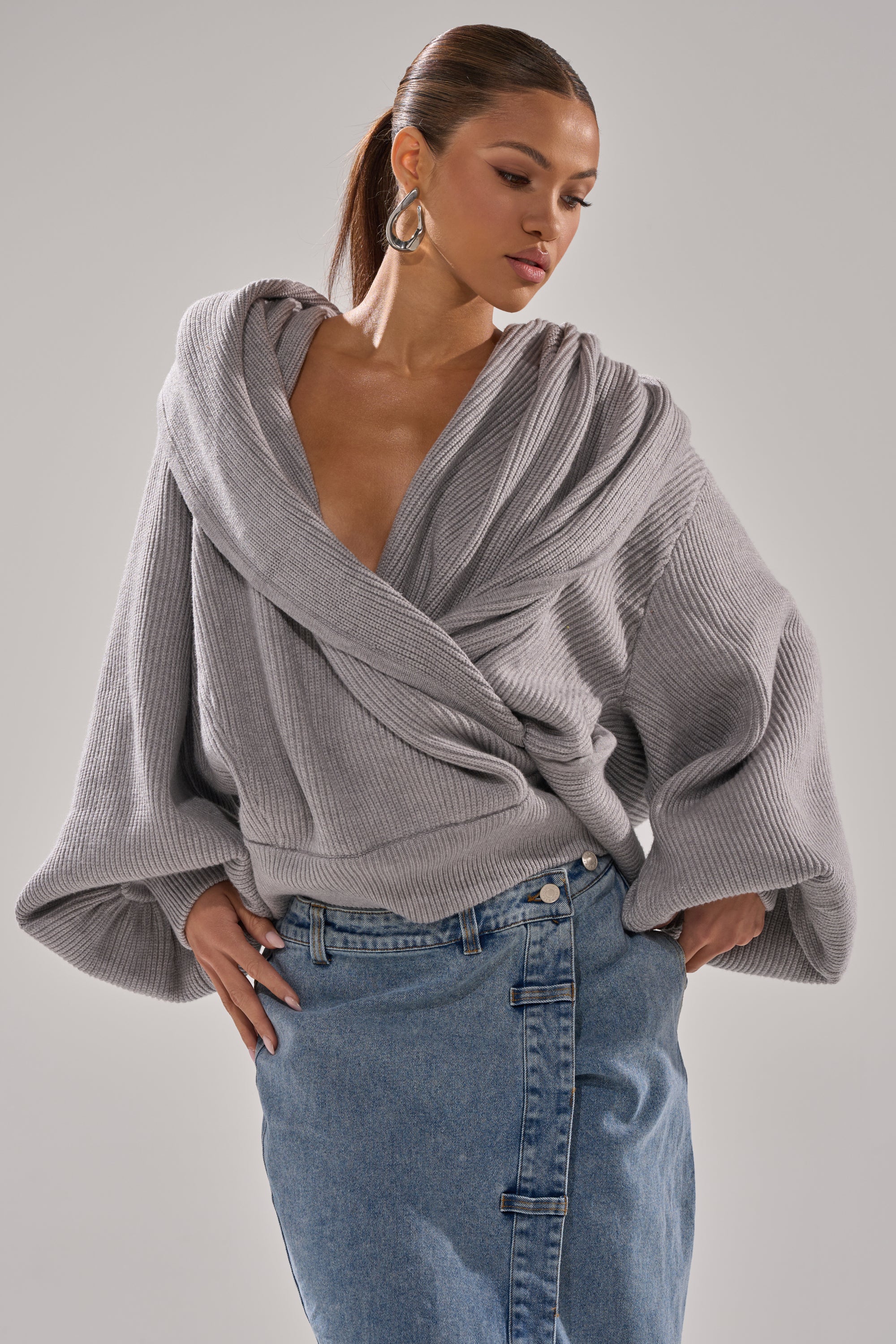 A woman models the CRAZY FOR YOU CROSS FRONT SWEATER in heather grey with wide sleeves and a deep neckline, paired with a high-waisted blue denim skirt, against a plain background. 1