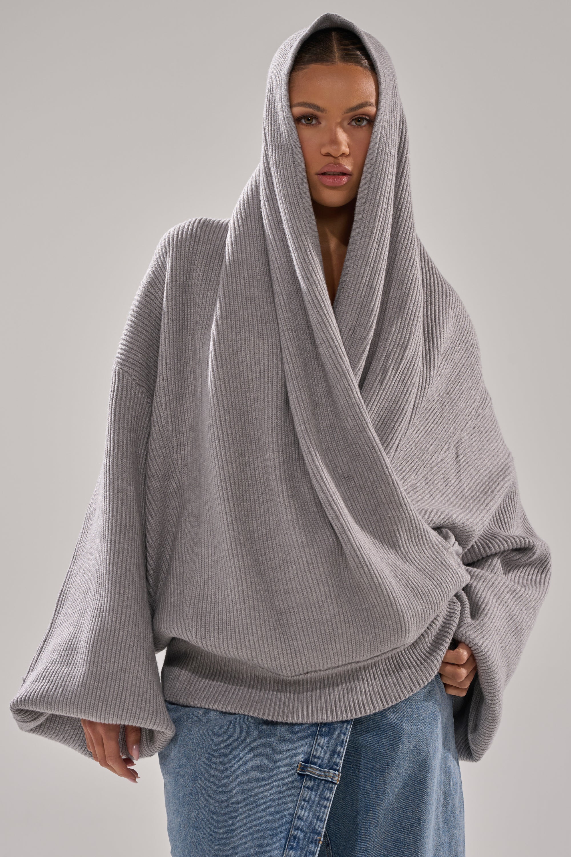 A woman wears the CRAZY FOR YOU CROSS FRONT SWEATER in heather grey, featuring wide sleeves and a hood, paired with blue jeans against a plain light gray background. 3