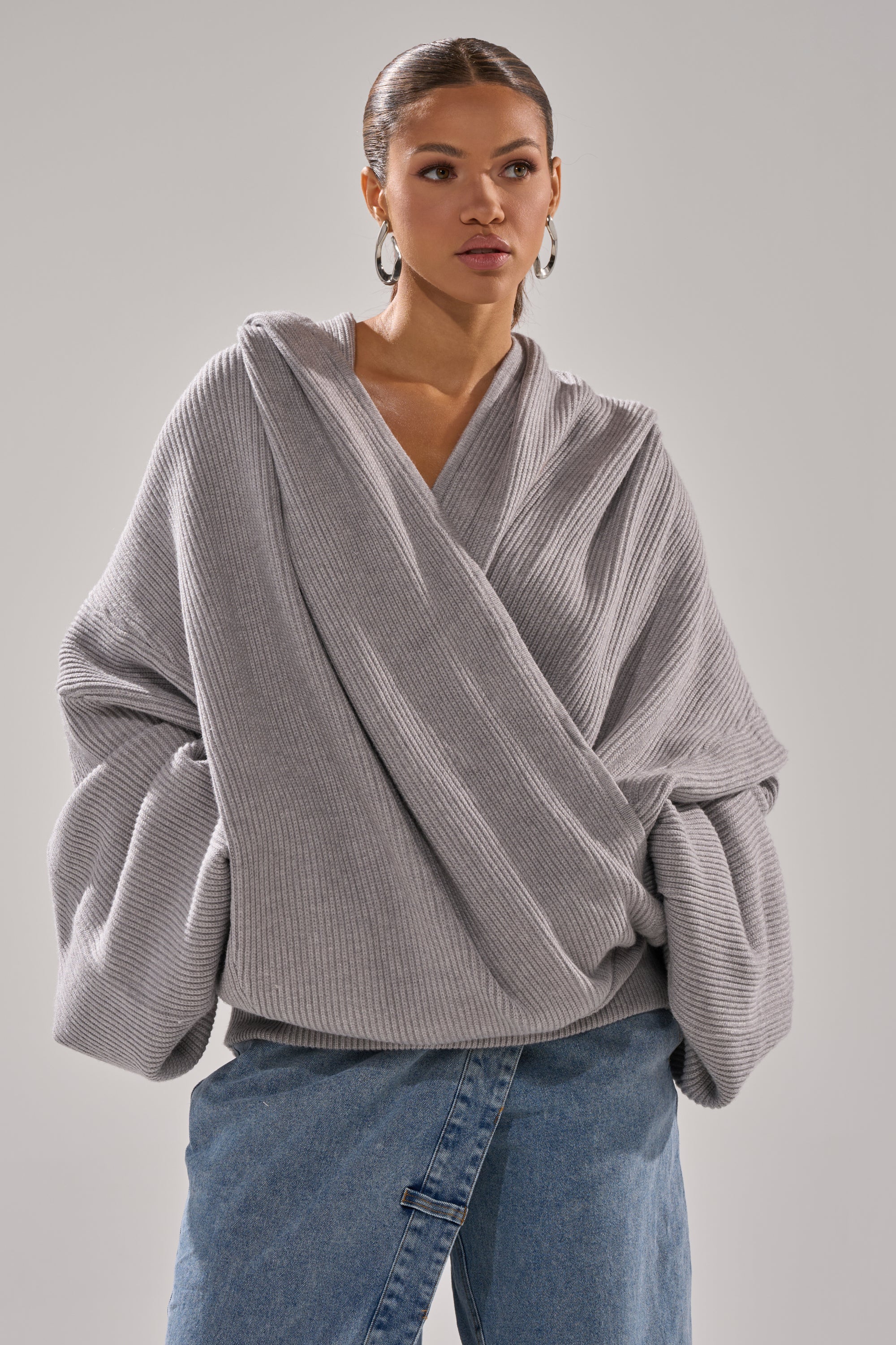 A person wears the CRAZY FOR YOU CROSS FRONT SWEATER, a loose heather grey top with wide sleeves and ribbed knit texture, paired with blue jeans and standing against a plain light gray background. 4