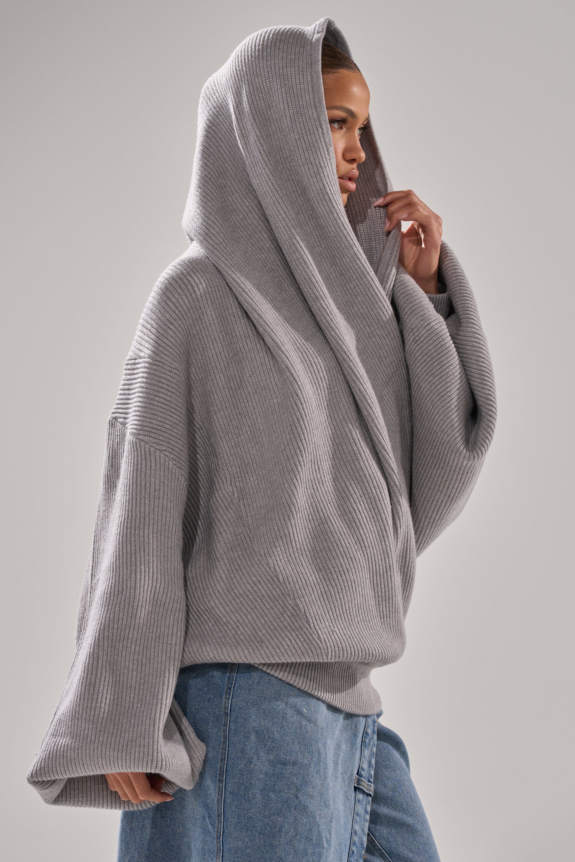 Model stands sideways in the CRAZY FOR YOU CROSS FRONT SWEATER, a loose, cozy heather grey top with a hood, paired with blue jeans, against a plain light background. One hand adjusts the hood. 6