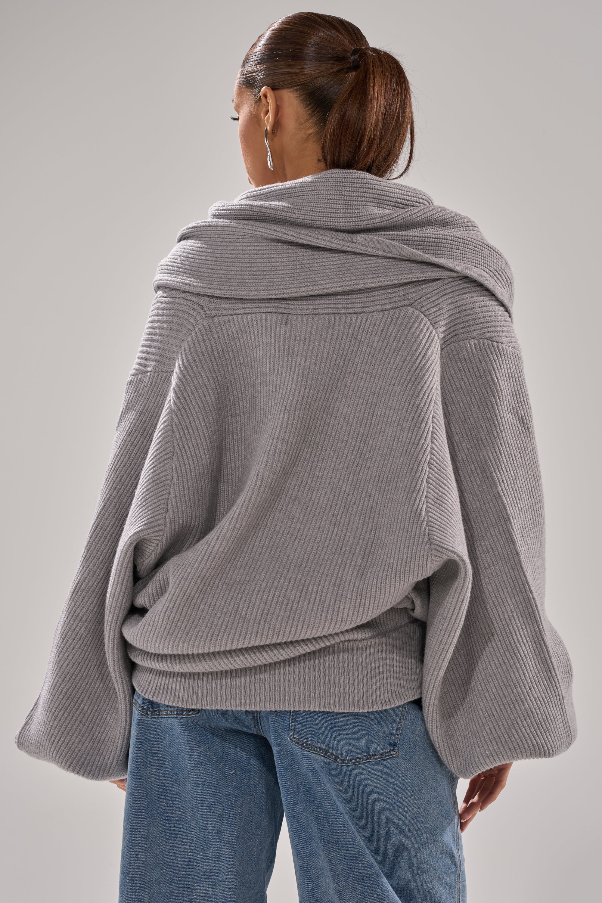 A person with long brown hair in a ponytail wears the CRAZY FOR YOU CROSS FRONT SWEATER, a loose-fitting heather grey top with a draped collar and wide sleeves, paired with blue jeans, seen from the back against a light gray background. 7