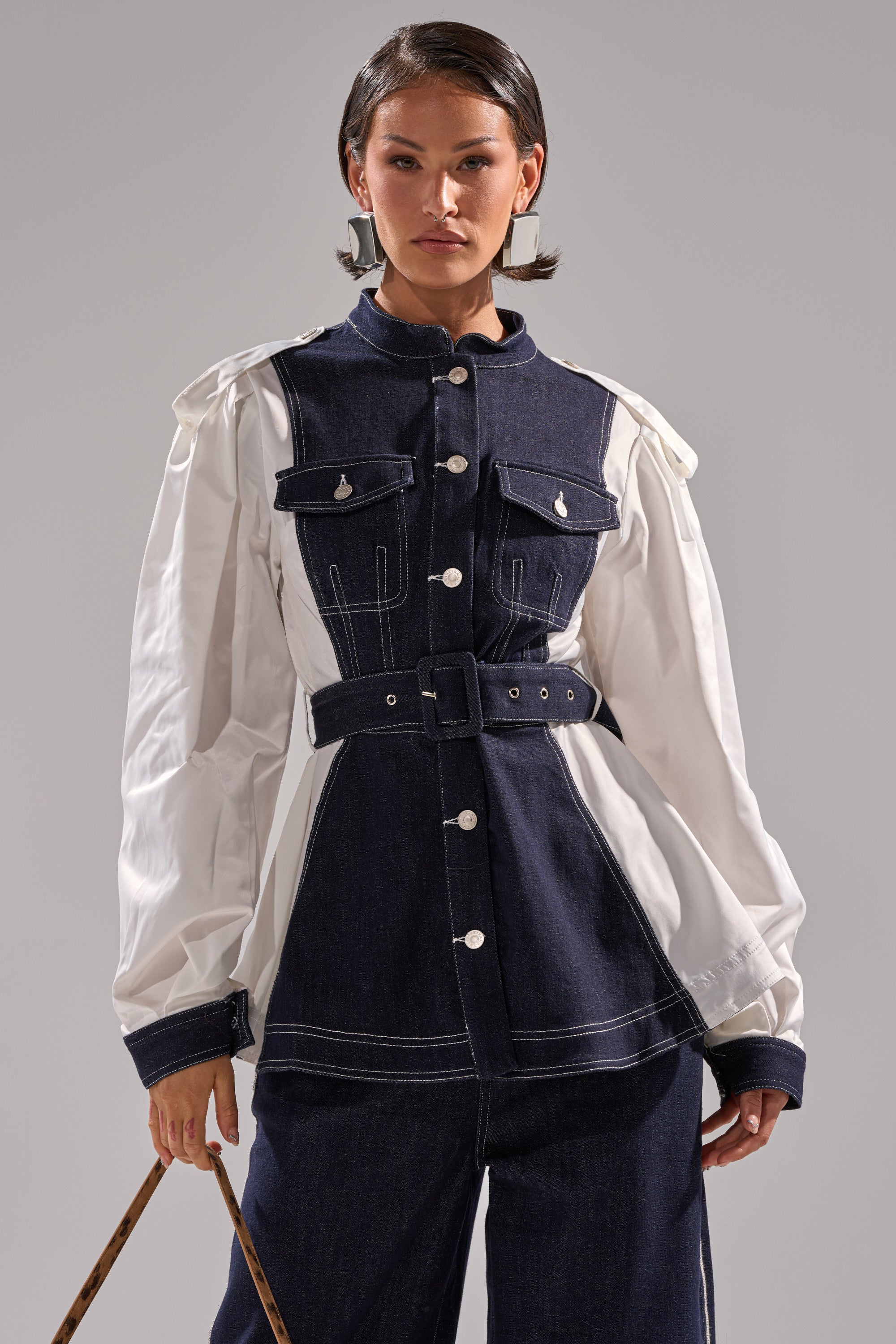 A woman stands confidently against a plain gray background, wearing the DIANA DENIM AND POPLIN BUTTON DOWN with navy denim panels, white sleeves, a belted waist, and matching navy pants.