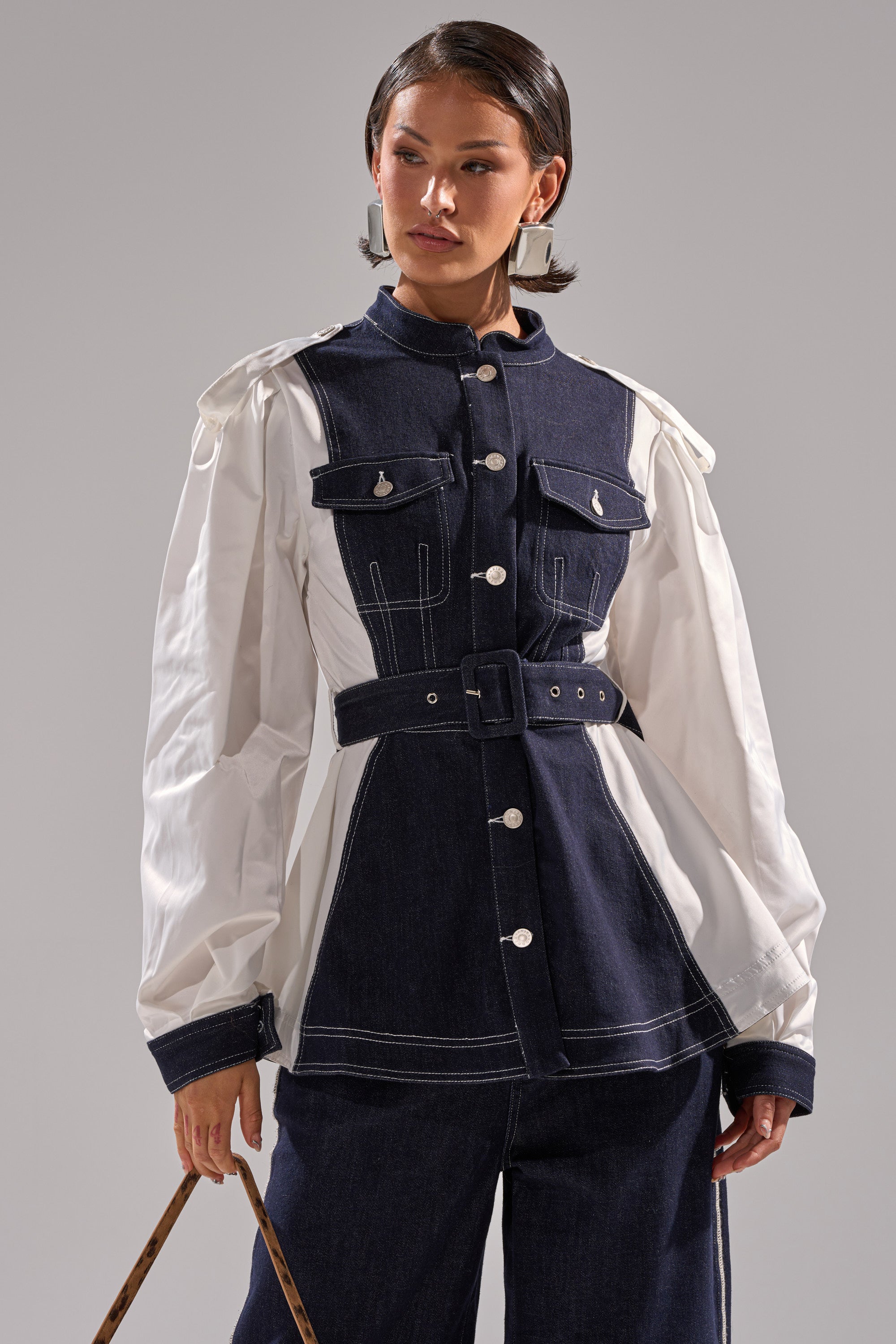 A woman stands against a plain gray background, wearing the DIANA DENIM AND POPLIN BUTTON DOWN—a stylish belted jacket with dark denim panels and white puffed sleeves—paired with matching dark denim pants.