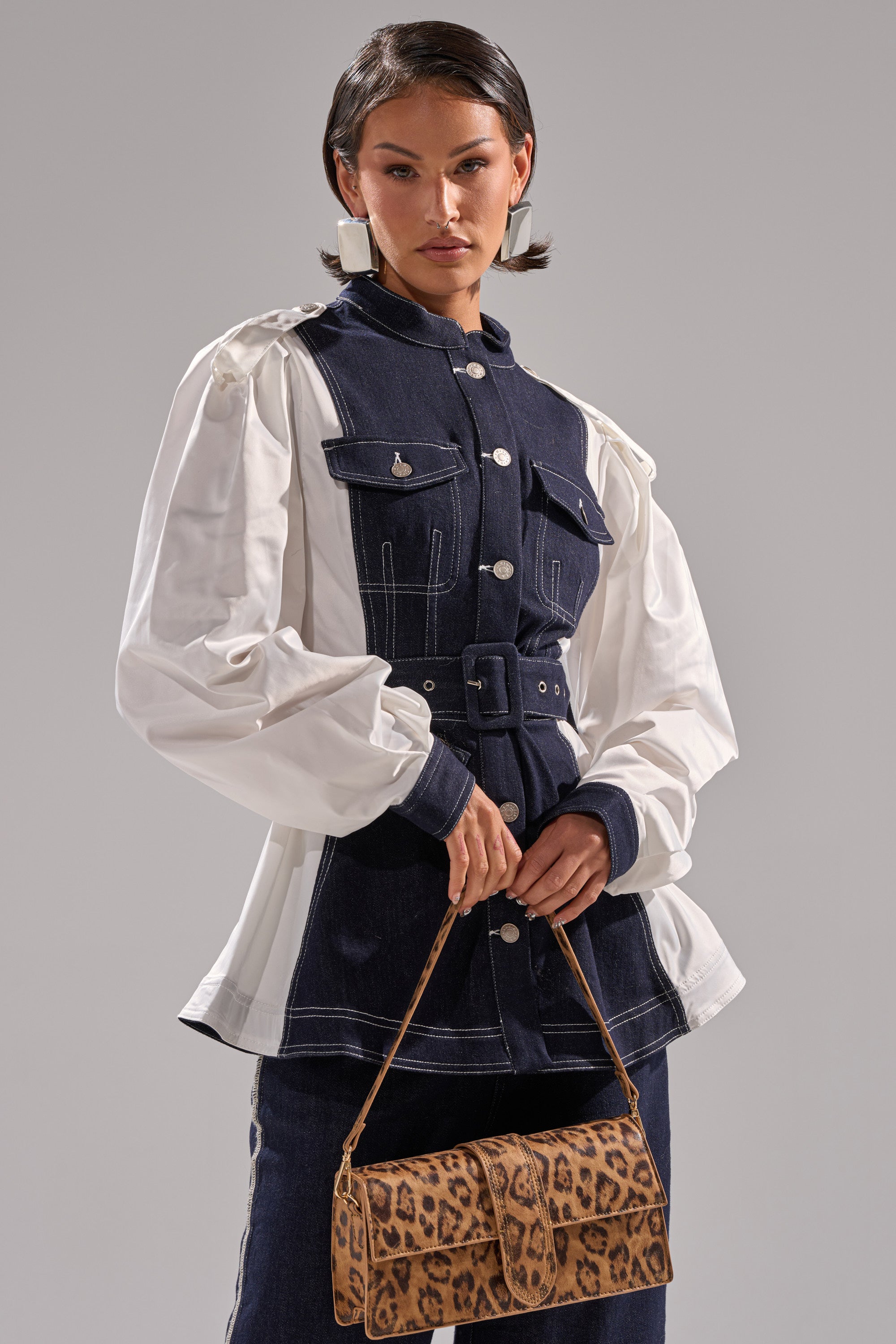A woman wears the DIANA DENIM AND POPLIN BUTTON DOWN, featuring a navy blue belted waist and white puffed sleeves, and holds a leopard-print handbag against a plain gray background.