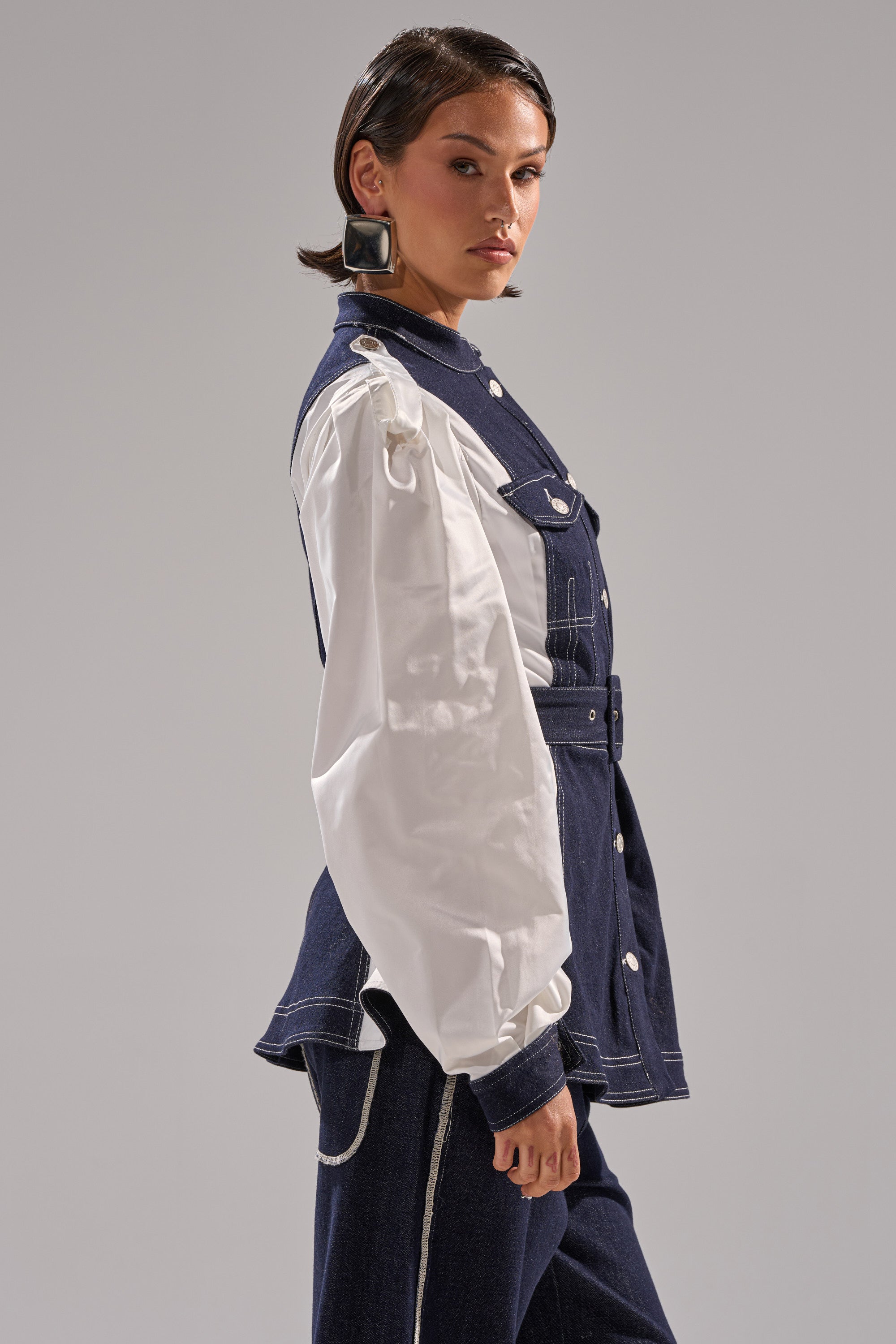 A woman wears the DIANA DENIM AND POPLIN BUTTON DOWN—a mixed-media jacket with a navy denim vest, white poplin sleeves, and white stitching—paired with matching navy pants and black square earrings, against a light gray background.