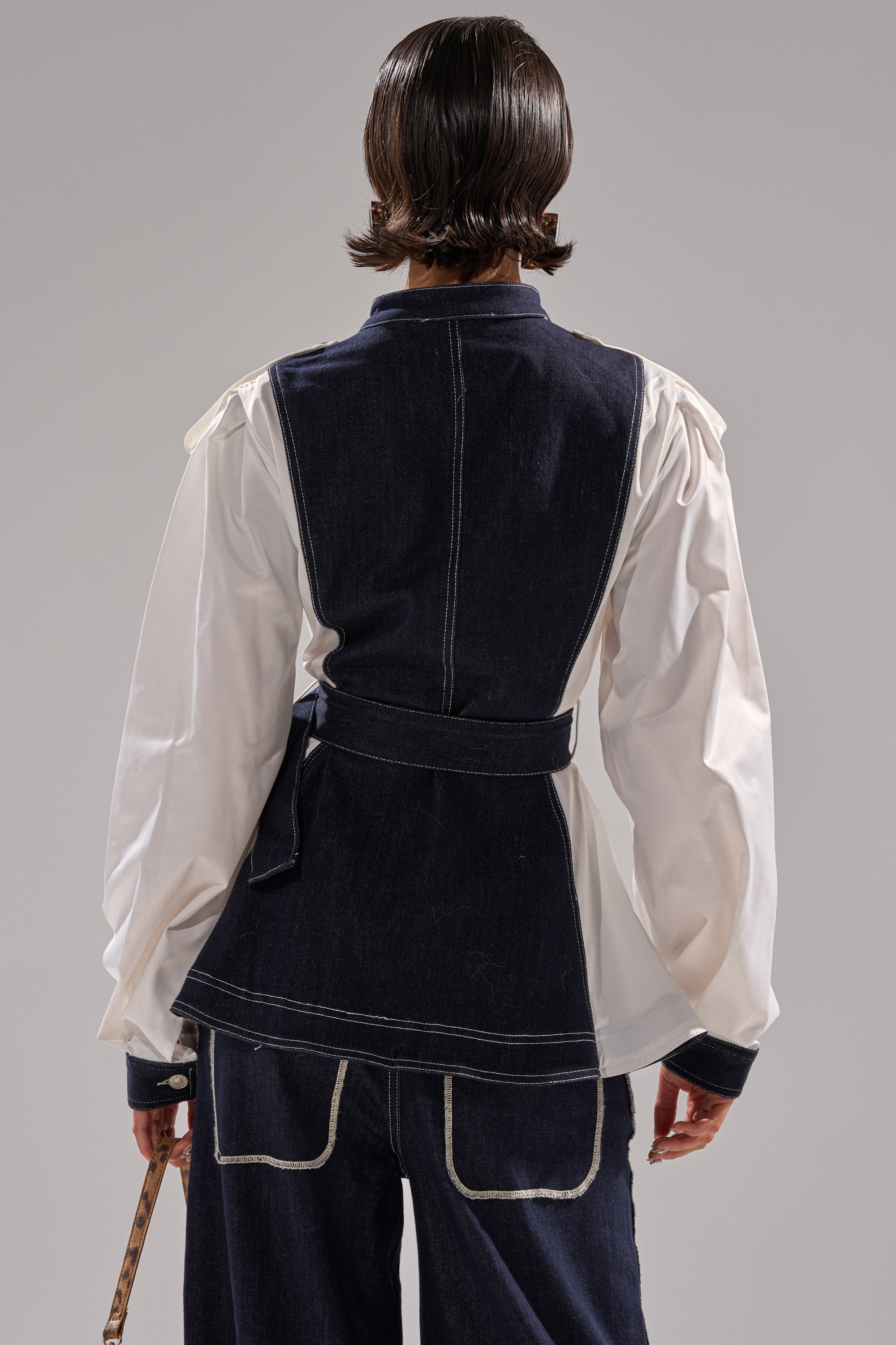 A person faces away, wearing the DIANA DENIM AND POPLIN BUTTON DOWN—a dark denim jacket with white stitching, belted waist, and white poplin sleeves—paired with matching denim pants. The background is plain and neutral.