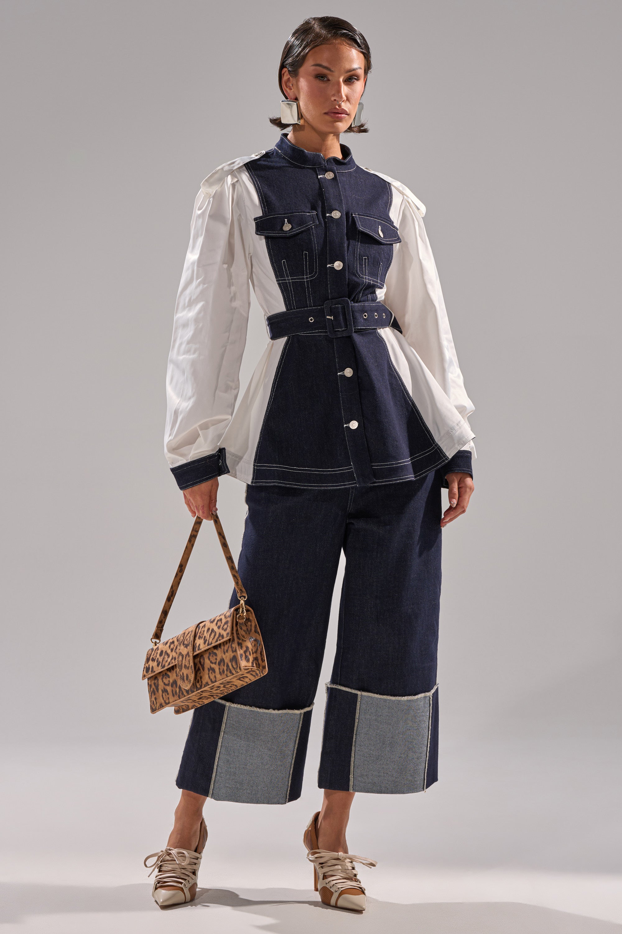 A woman wears the DIANA DENIM AND POPLIN BUTTON DOWN with a belted waist and white sleeves, styled with wide-leg cropped denim pants, a leopard-print handbag, beige lace-up shoes, and loose waves against a plain background.