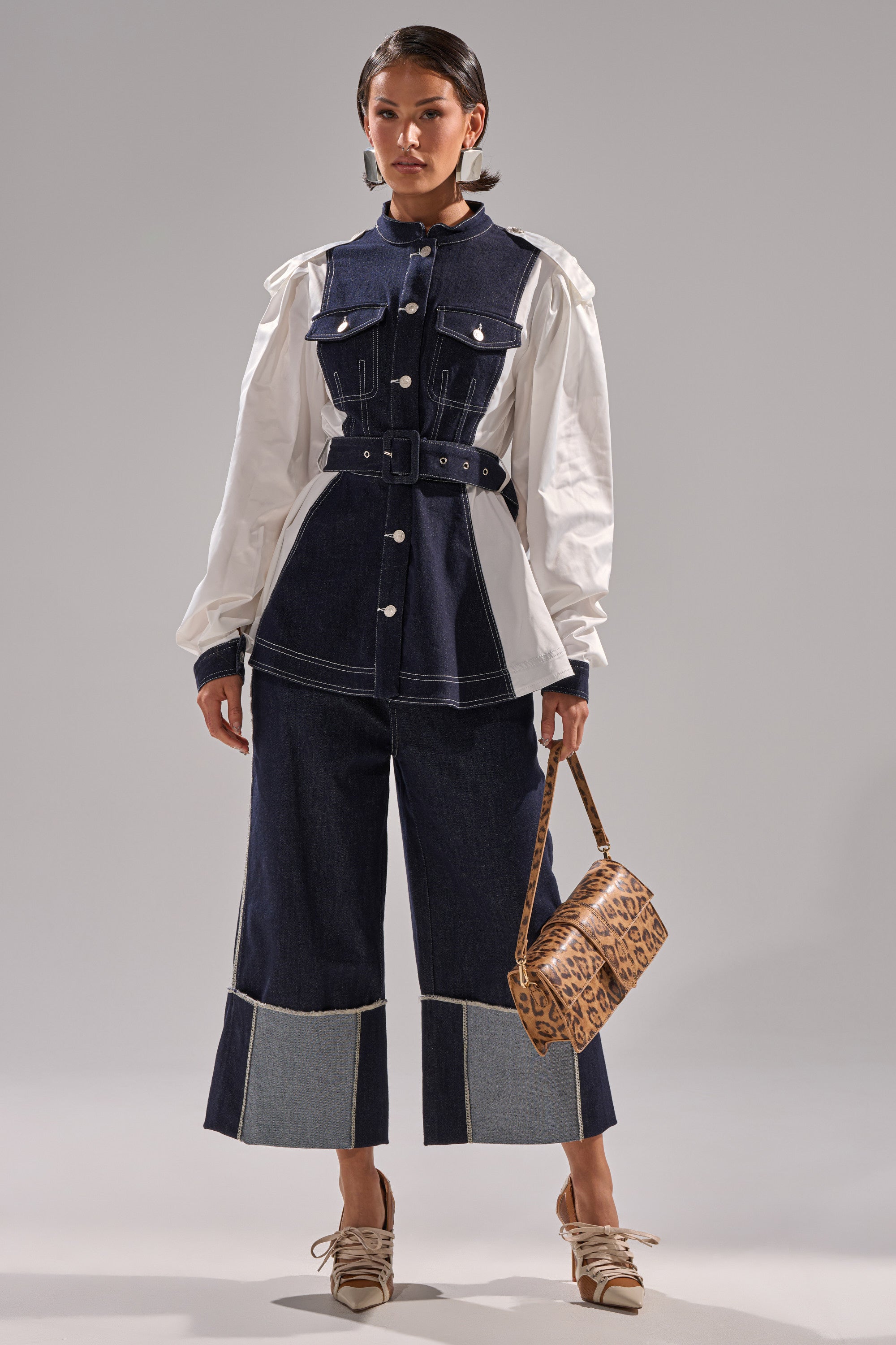 A woman wears the DIANA DENIM AND POPLIN BUTTON DOWN—a mixed-media jacket with a belted waist and white puffed sleeves—paired with wide-leg denim pants, tan lace-up shoes, and a small leopard-print handbag.