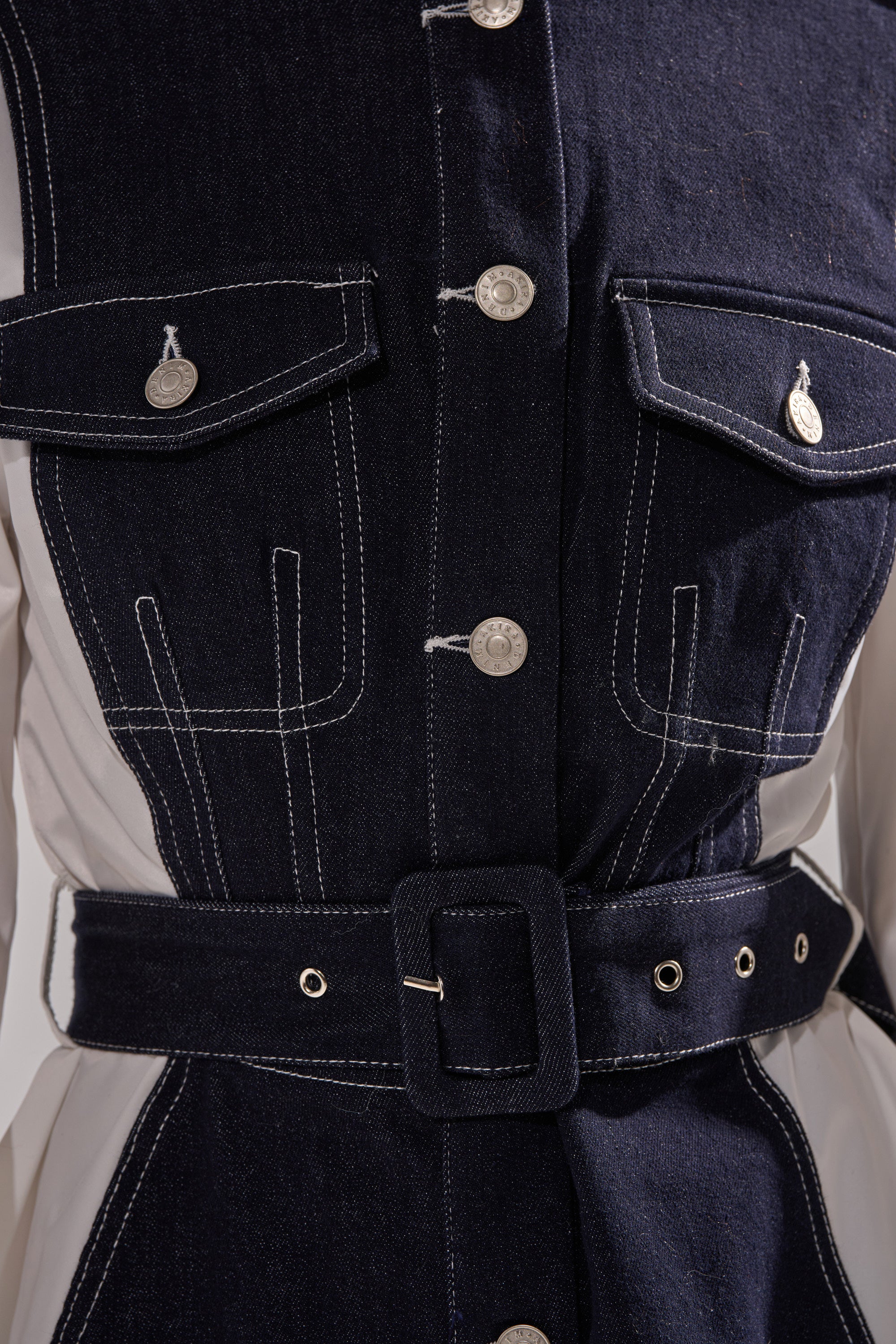 Close-up of the DIANA DENIM AND POPLIN BUTTON DOWN, featuring dark denim, white stitching, silver buttons, flap chest pockets, and a matching denim belt with a rectangular buckle, styled over a light-colored layer.