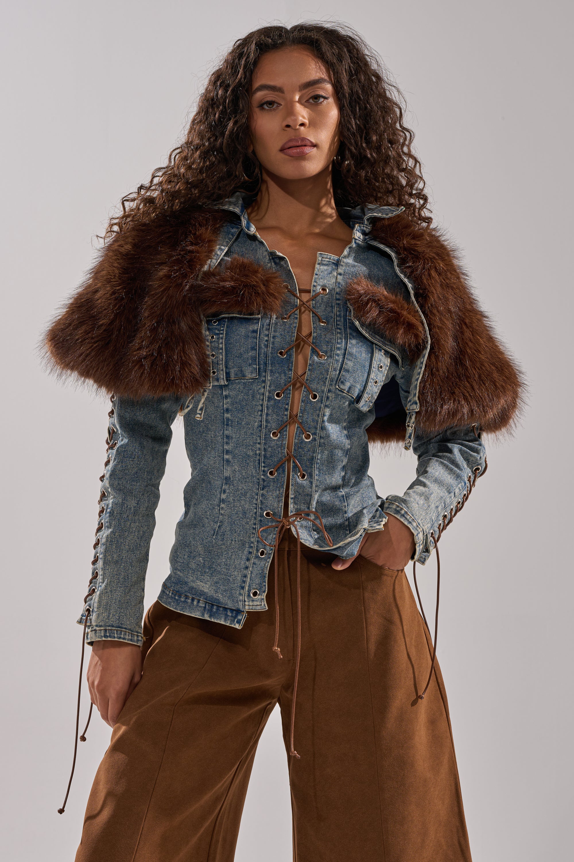A woman with long curly hair models the TRY ME BABY LACE UP DENIM TOP, featuring lace-up details and brown faux fur shoulders, paired with wide-leg brown trousers as she poses confidently against a plain background.