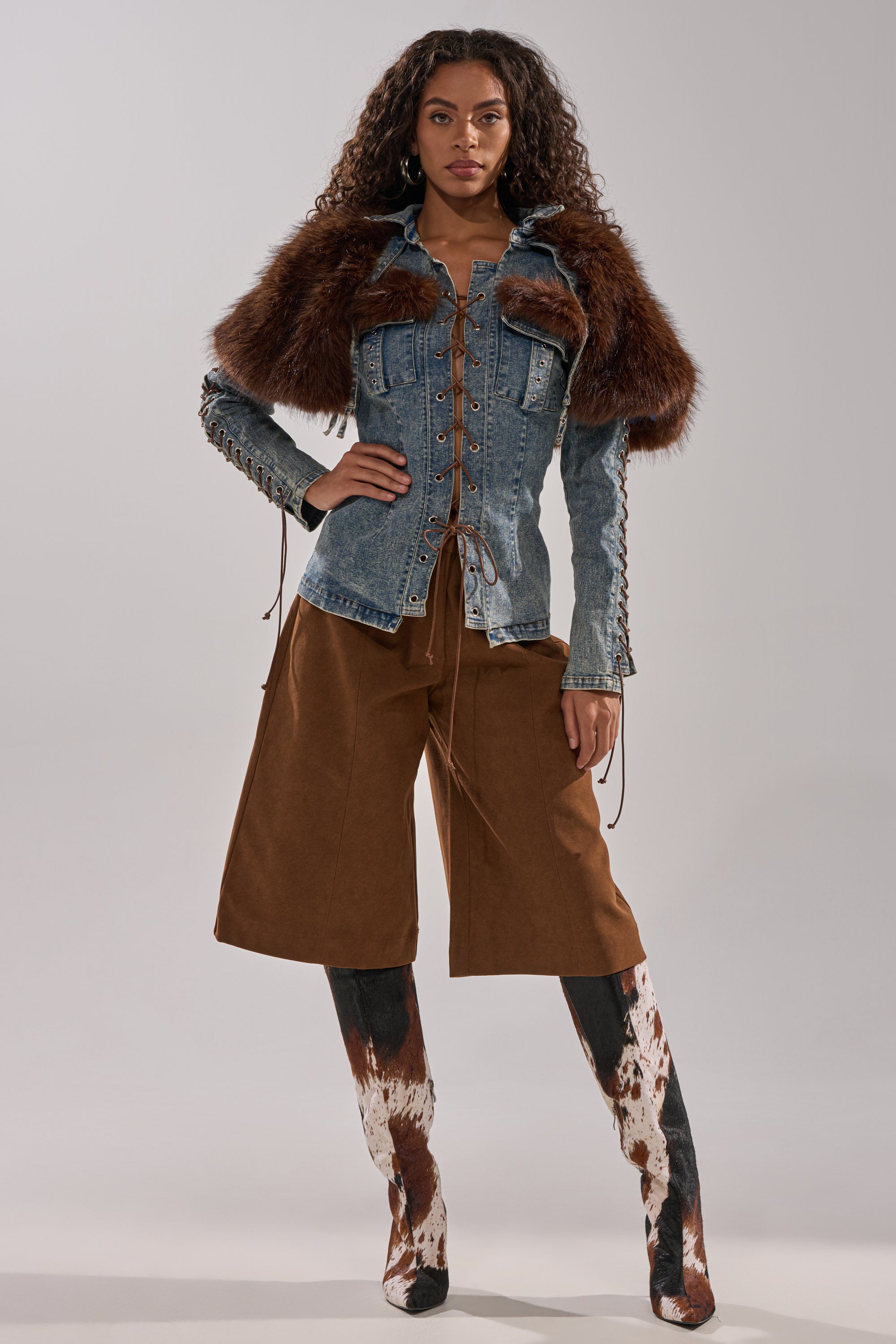 A woman stands confidently in the TRY ME BABY LACE UP DENIM TOP with faux fur shoulder panels, wide brown culottes, and knee-high cow-print boots against a plain gray background.