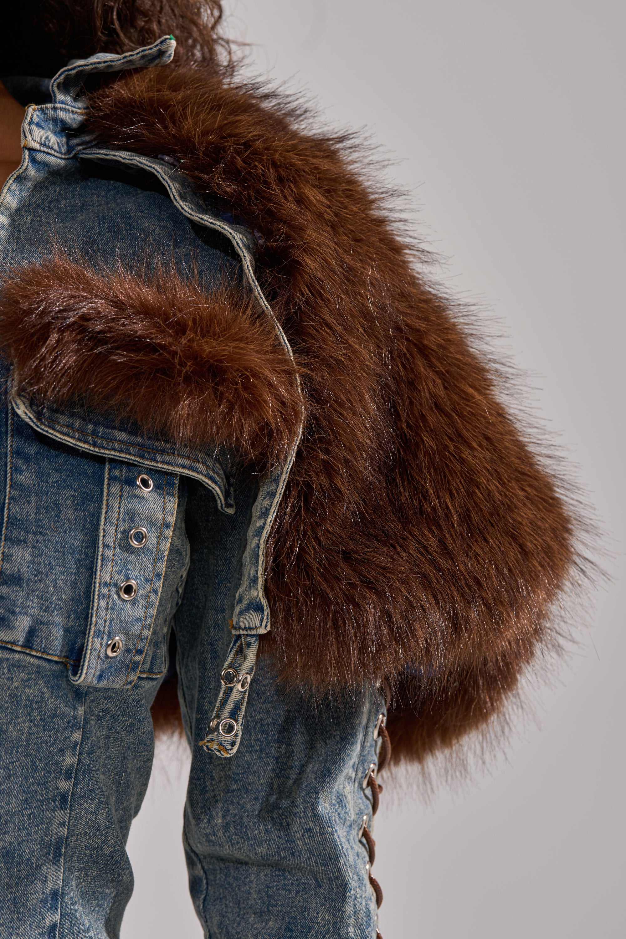 Wearing the TRY ME BABY LACE UP DENIM TOP with metallic eyelets and lace-up details, styled with a large, fluffy brown faux fur panel draped over one shoulder.