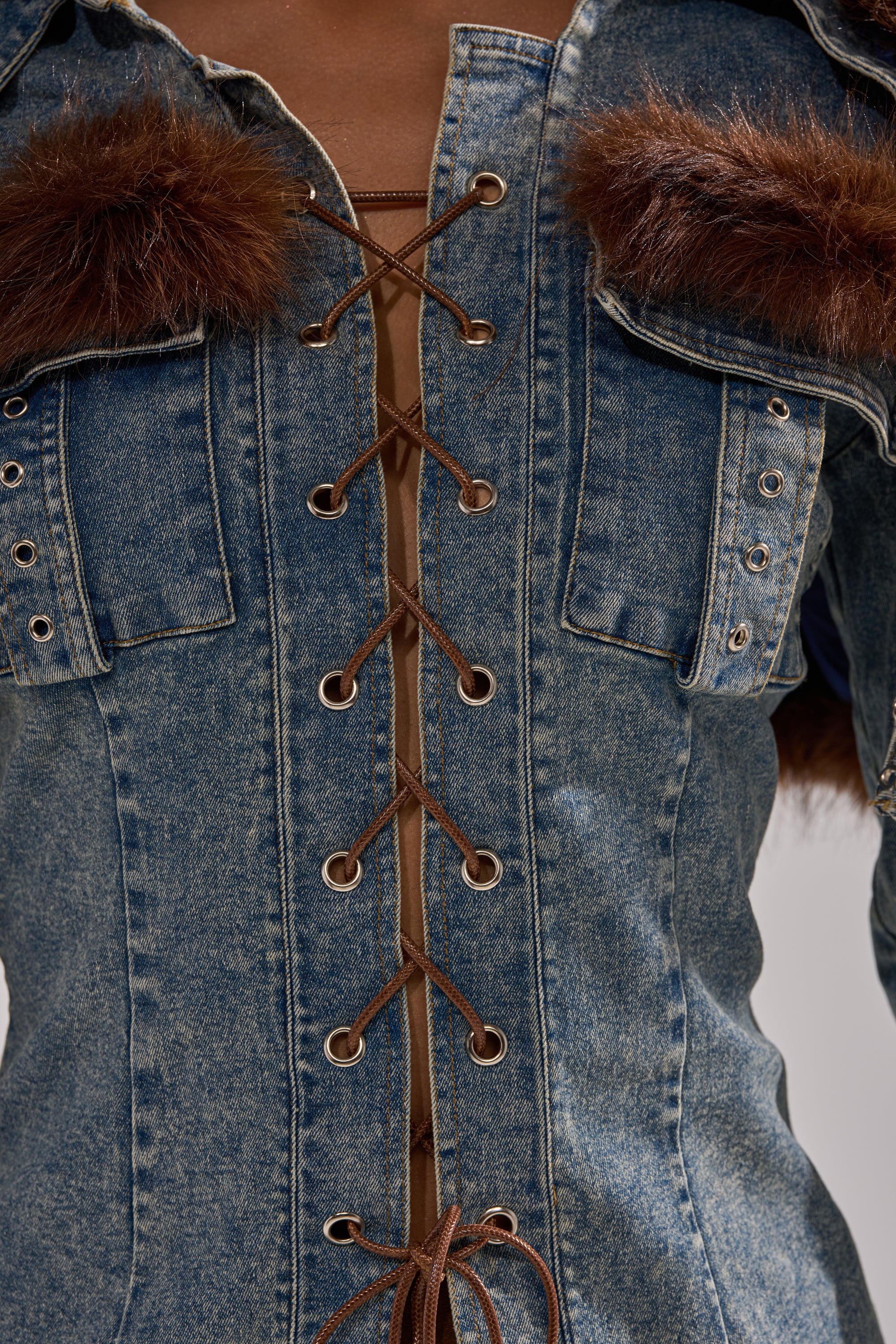 Close-up of the TRY ME BABY LACE UP DENIM TOP, featuring corset-inspired brown lace-up detailing, faux fur on chest pockets, metal eyelets, and decorative stitching. The top reveals skin with its stylish open front design.