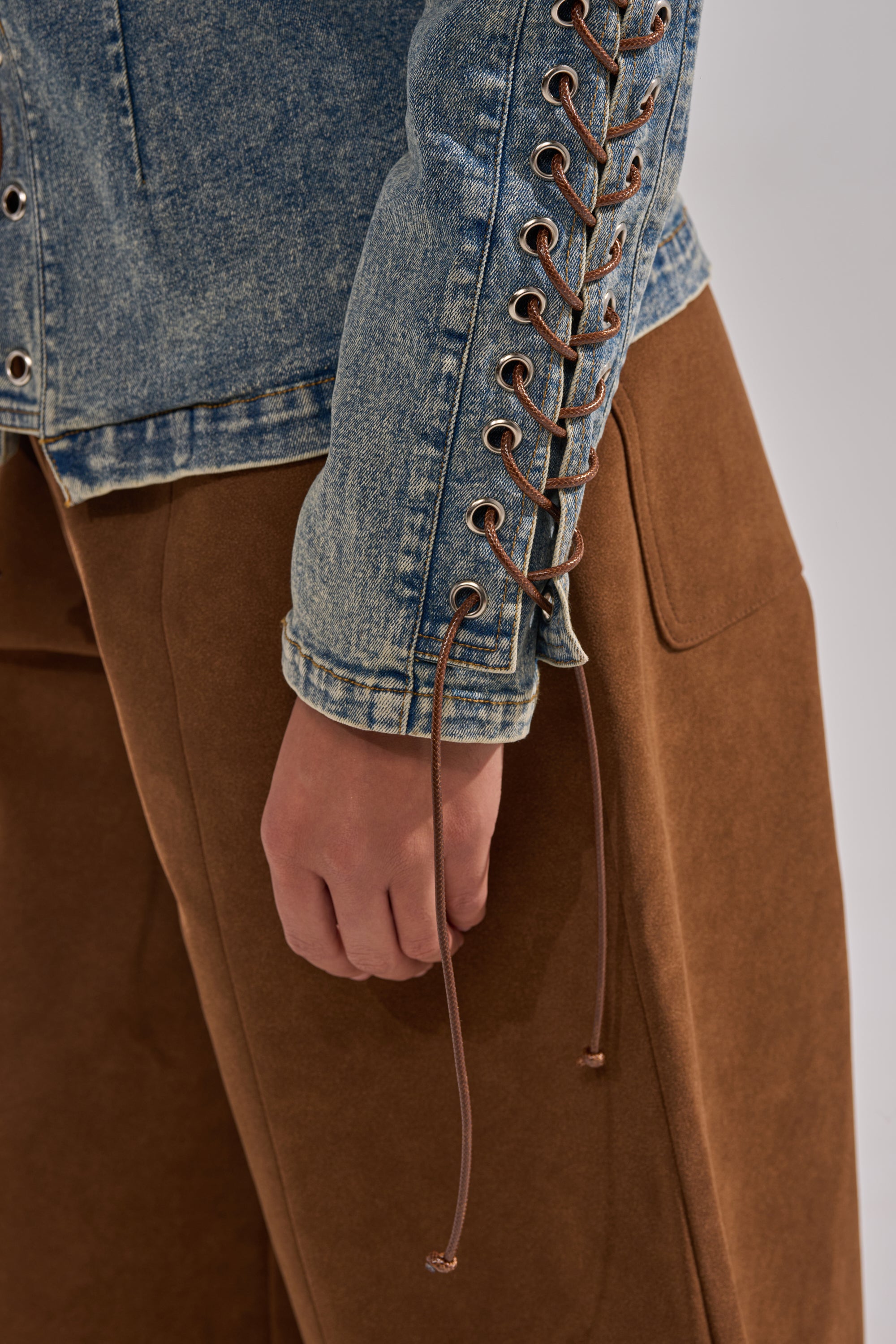 A person wearing brown pants and the TRY ME BABY LACE UP DENIM TOP, a corset-inspired blue denim jacket with lace-up sleeve detailing, shown from the waist down with their hand relaxed at their side.