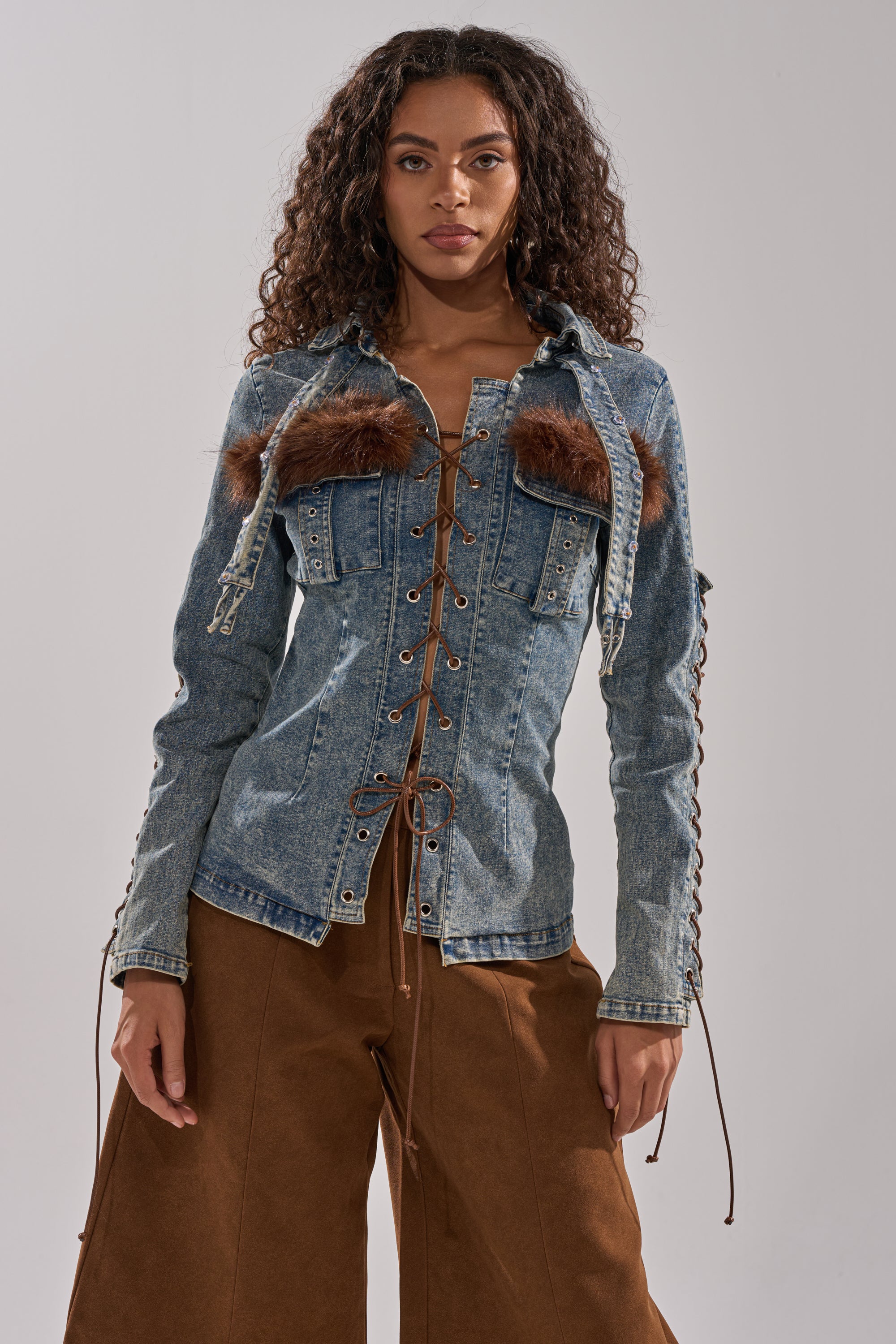 A woman with long curly hair wears the TRY ME BABY LACE UP DENIM TOP featuring corset-inspired lace-up details and faux fur panels, paired with brown wide-leg pants, standing against a plain light background.