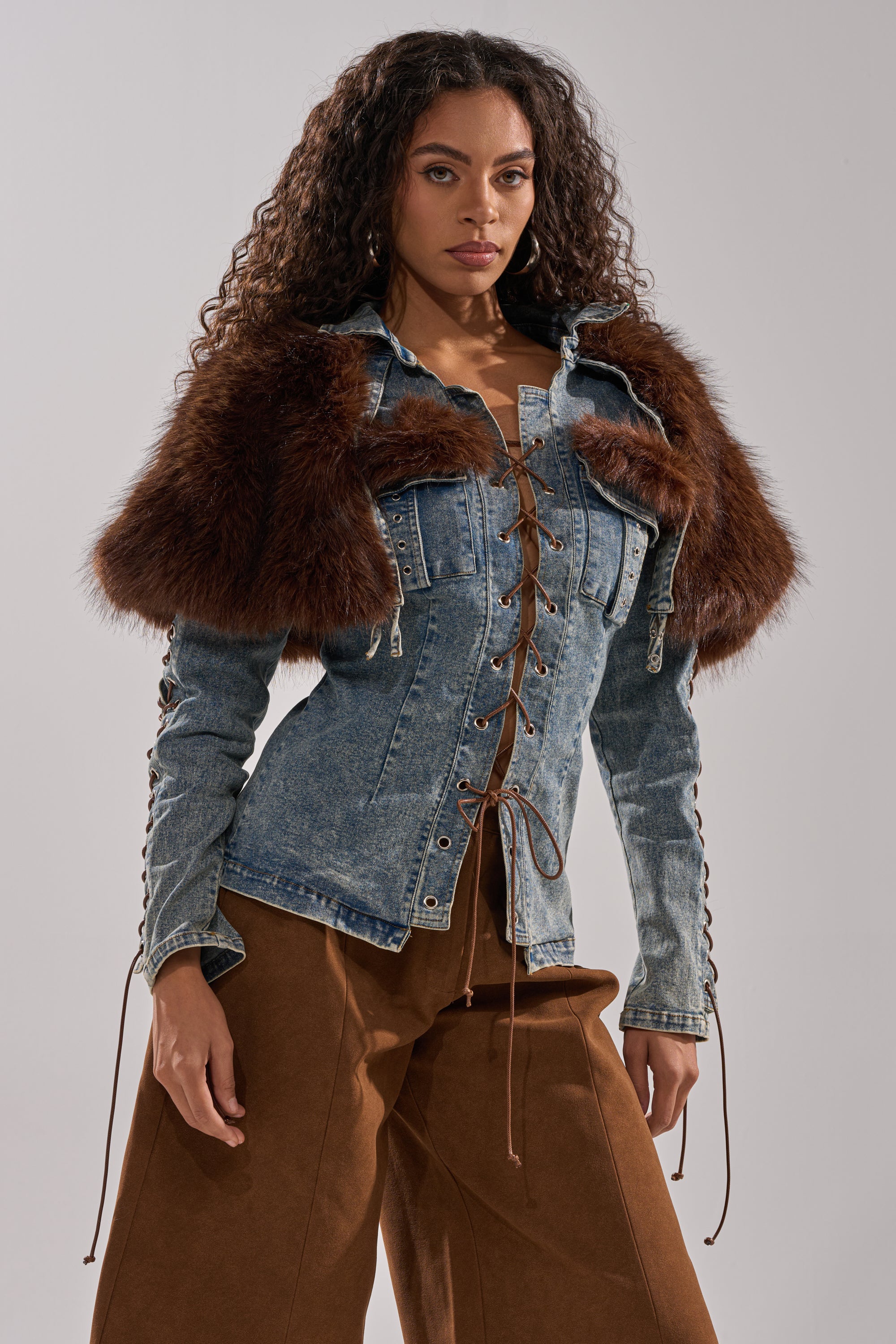 A woman with long curly hair wears the TRY ME BABY LACE UP DENIM TOP, a corset-inspired piece with a faux fur panel across the shoulders, paired with brown wide-leg pants against a plain light background.
